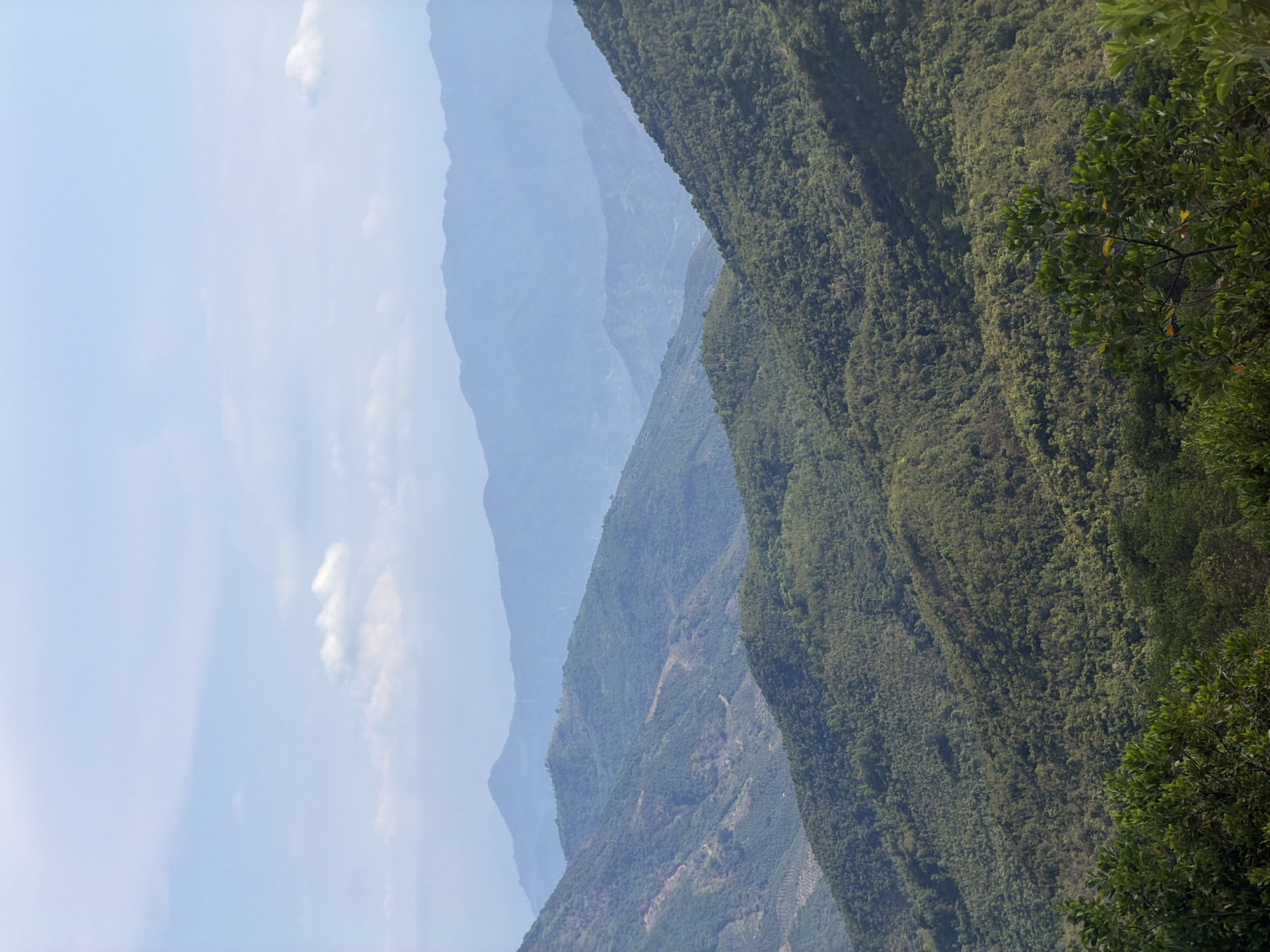 The image presents a view of layered mountains in a region of Nam Nha Trang Ward, Vietnam. The landscape is dominated by mountain ranges, with the closest one covered in dense green vegetation, including trees. Further back, the mountains gradually fade into a blue-grey hue, indicating distance and atmospheric perspective. The sky is light blue, with the edges of clouds visible in the left. The photo seems to be taken from an elevated position, providing a panoramic perspective of the natural landscape.