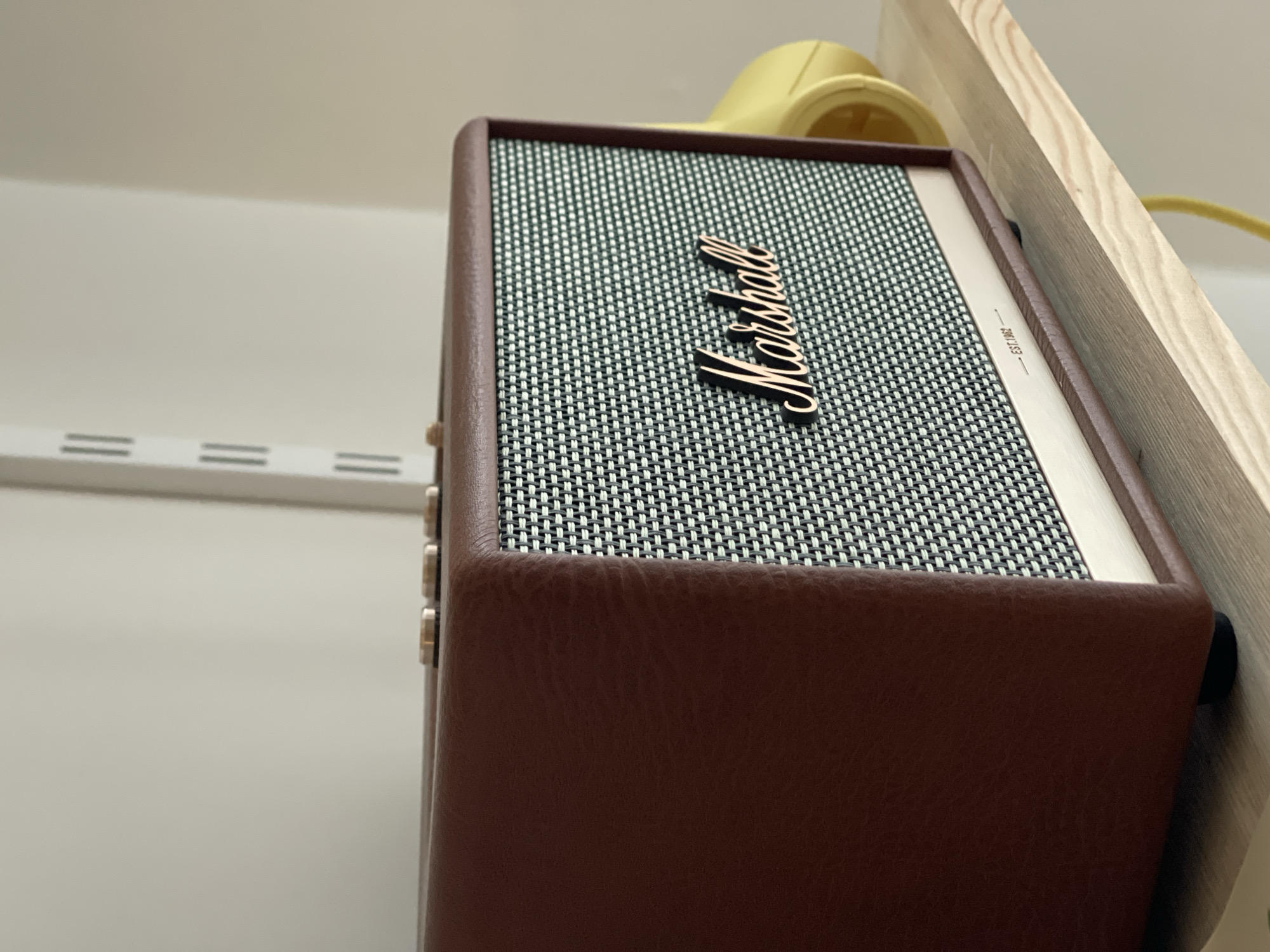 The image shows a brown Marshall Kilburn II speaker sitting on a light wooden shelf. The speaker has the Marshall logo in gold lettering on a patterned speaker grill. The speaker is located near a white wall with a metal rack with horizontal slots. A yellow object with a visible cord is situated behind the speaker. There is no information to confirm that this picture was taken in Izhevsk, Russia.