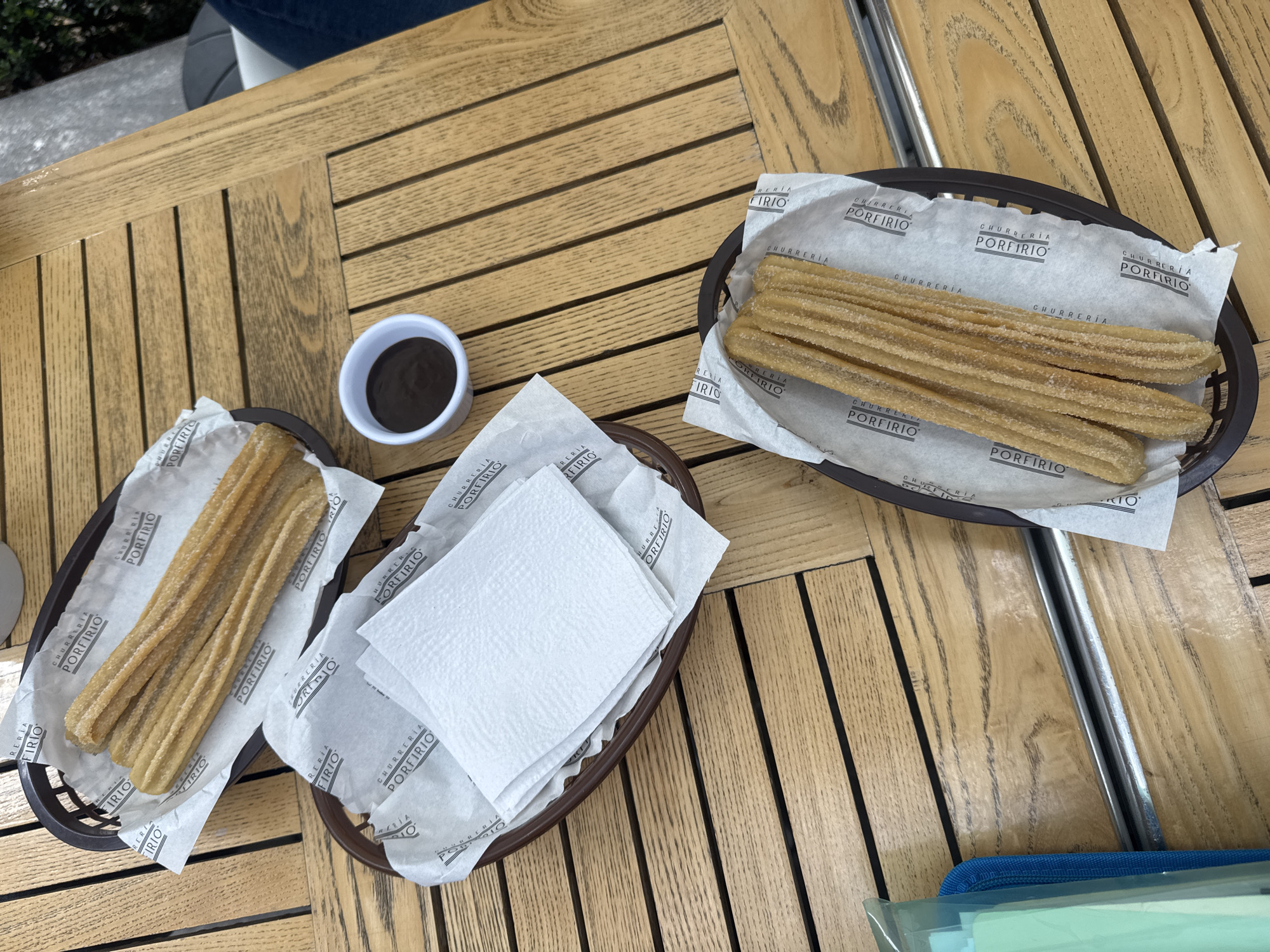 The image presents an overhead view of a wooden slatted table, likely at an outdoor cafe or restaurant, offering a snack of churros. Two brown, oval-shaped baskets each contain several long, sugar-coated churros, arranged neatly on white paper liners. A third, smaller brown basket, also lined with similar branded paper, holds a stack of white paper napkins. Between the baskets of churros, a small white cup is filled with dark chocolate dipping sauce.
The paper liners prominently display the text "CHURRERÍA PORFIRIO" and "PORFIRIO," indicating the establishment's name. The bright, natural lighting suggests it is daytime. The overall scene depicts a casual and inviting atmosphere for enjoying churros and chocolate, a common and beloved treat, particularly in Mexico City, Mexico.