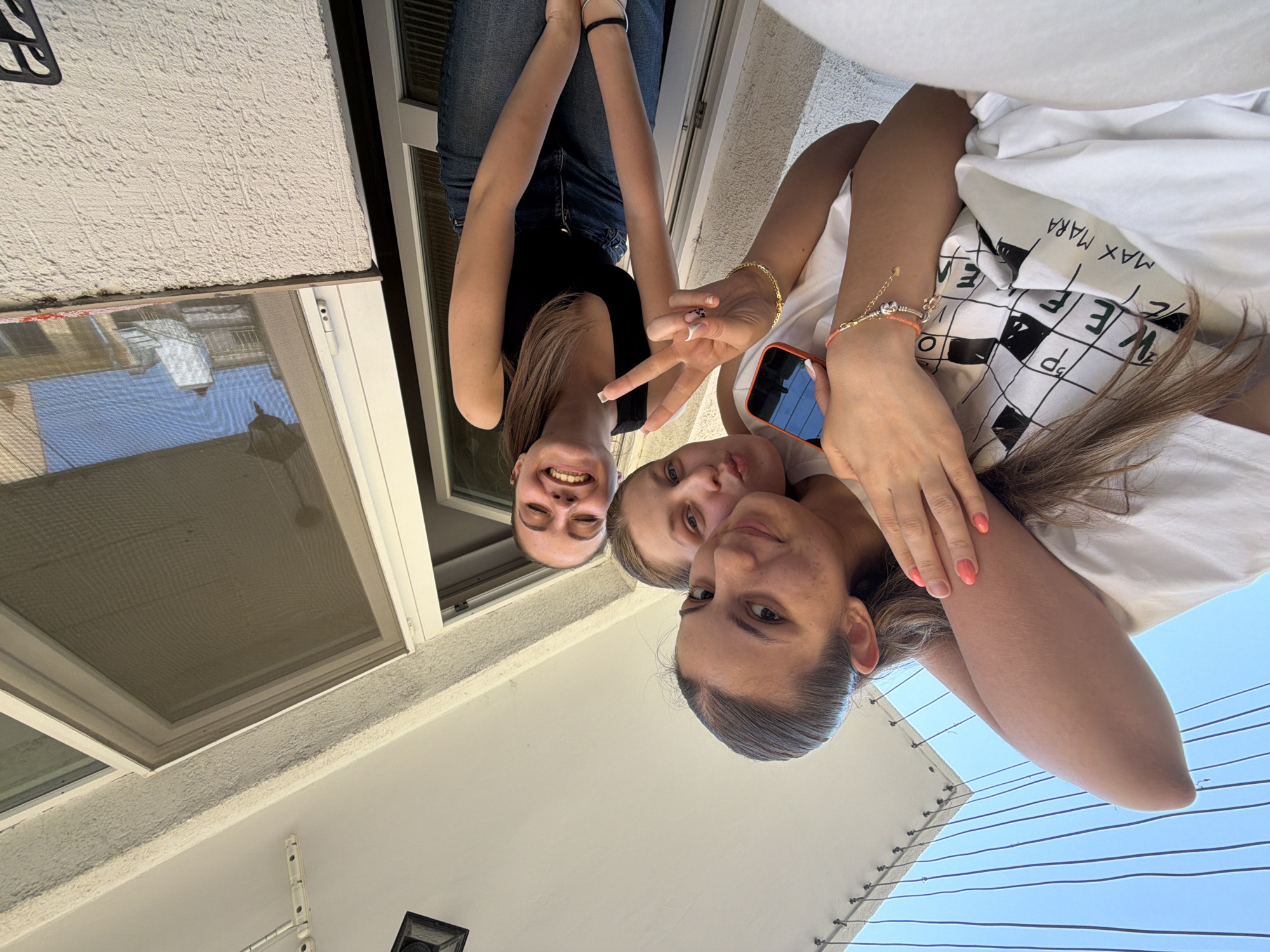 The image, originally oriented with subjects on their sides, depicts three young women in a bright outdoor setting, likely a balcony or patio in Burgas, Bulgaria. They are smiling towards the camera, suggesting a happy and casual group photo.
The woman on the left, with long brown hair, wears a black sleeveless top and blue jeans. She smiles broadly and forms a peace sign with her right hand, adorned with bracelets. The middle woman, with lighter hair, is partially visible, also smiling at the camera. The woman on the right, in the foreground, has dark hair pulled back and is dressed in a white t-shirt. She holds a smartphone with a bright orange case in her left hand, its screen reflecting the blue sky. Her right wrist features multiple bracelets, including one with coral-colored beads, and her fingernails are painted a matching coral-pink.
The background features a textured, light-colored stucco wall to the top and left, which includes a white-framed window with a mesh screen. A black patterned mat is visible on the ground near the wall. In the lower right background, a clear blue sky is visible, traversed by several thin, dark utility wires. Part of an outdoor light fixture or security camera is also present. The bright, even illumination indicates a clear, sunny day. The white t-shirt worn by the foreground woman has visible text, "MAX MARA", and a partial crossword-like pattern with letters such as "W E P d p".