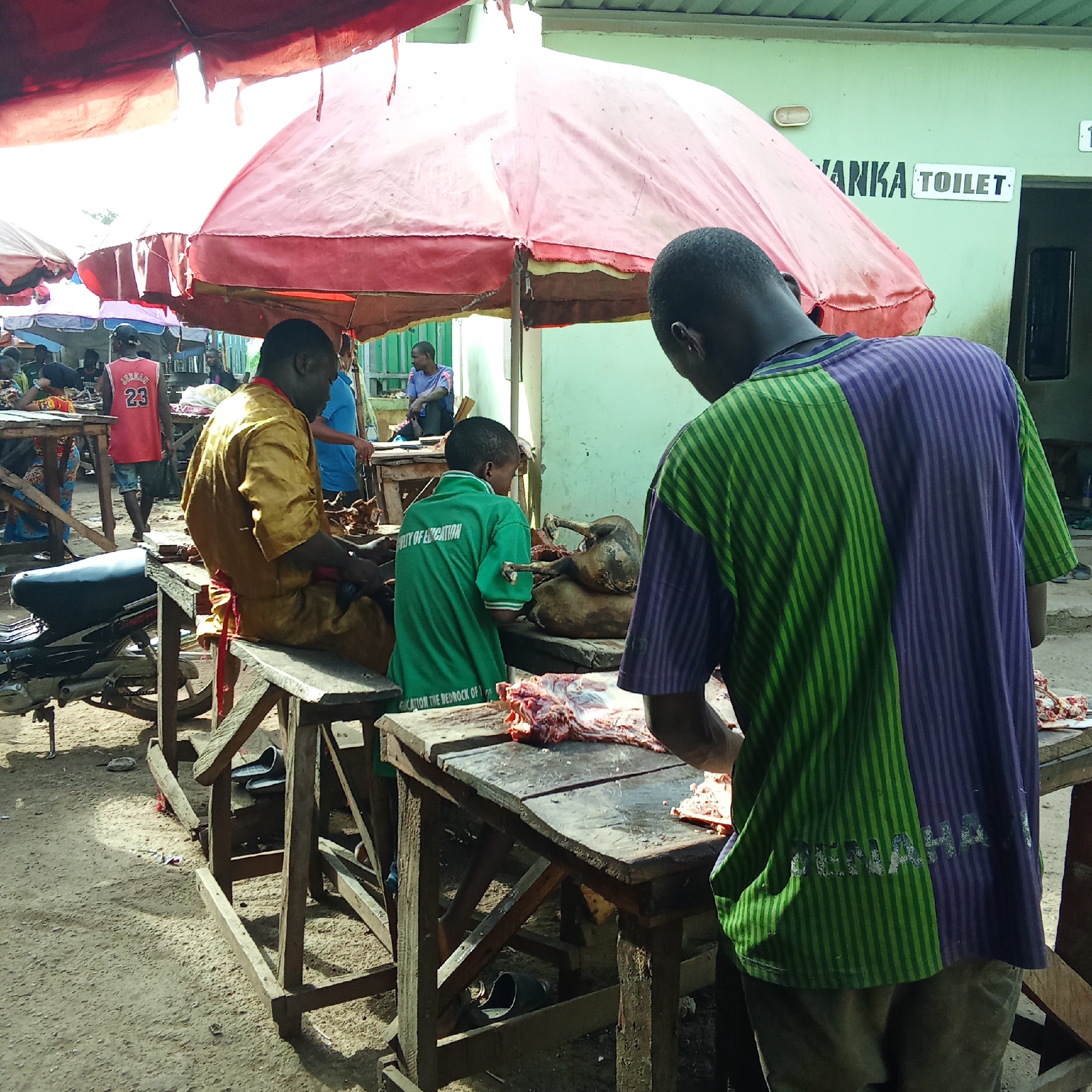 An outdoor market scene in Akwanga, Nigeria, shows several individuals engaged in meat preparation under large red awnings providing shade. The bright daylight suggests it is midday under sunny conditions.
In the foreground, a man with his back to the camera wears a distinctive green and purple striped shirt, featuring a faint logo that appears to read "RENAHA" on the lower back. He is standing at a wooden table, seemingly cutting raw meat laid out before him. To his left, a young boy in a green t-shirt, which reads "FACULTY OF EDUCATION" and "CATION THE BEDROCK OF G" (likely part of "EDUCATION THE BEDROCK OF GROWTH"), is also focused on meat on an adjacent table. Further left, a man in a golden-yellow robe is seated on a wooden bench, engaged in similar work. On their tables, various cuts of meat are visible, including what looks like a partially processed animal carcass.
In the mid-ground, a man wearing a red "Jordan 23" jersey stands near other market stalls. A black motorbike is parked beneath one of the wooden tables. The ground is dusty dirt. In the background, a light green building is visible, bearing a sign that clearly states "ANKA TOILET". The overall impression is a bustling local market environment where butchery is taking place.