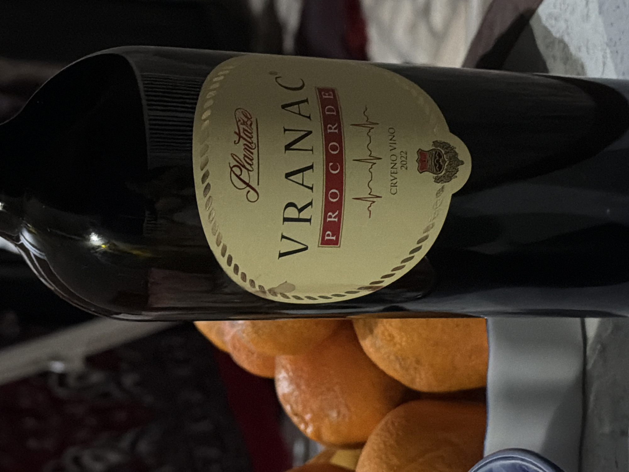 A horizontally oriented photograph depicts a bottle of "VRANAC PRO CORDE" red wine from Plantaze, specifically the 2022 vintage, positioned over a pile of oranges. The label is visible, including the "CRVENO VINO 2022" designation and a graphic resembling an electrocardiogram. The wine bottle is dark and glossy, contrasting with the beige label. The oranges below are a mix of smooth and textured surfaces. The background is blurry, containing indistinct shapes and colors, including what appears to be patterned fabric. Location in Krašići, Montenegro, cannot be confirmed from the image.