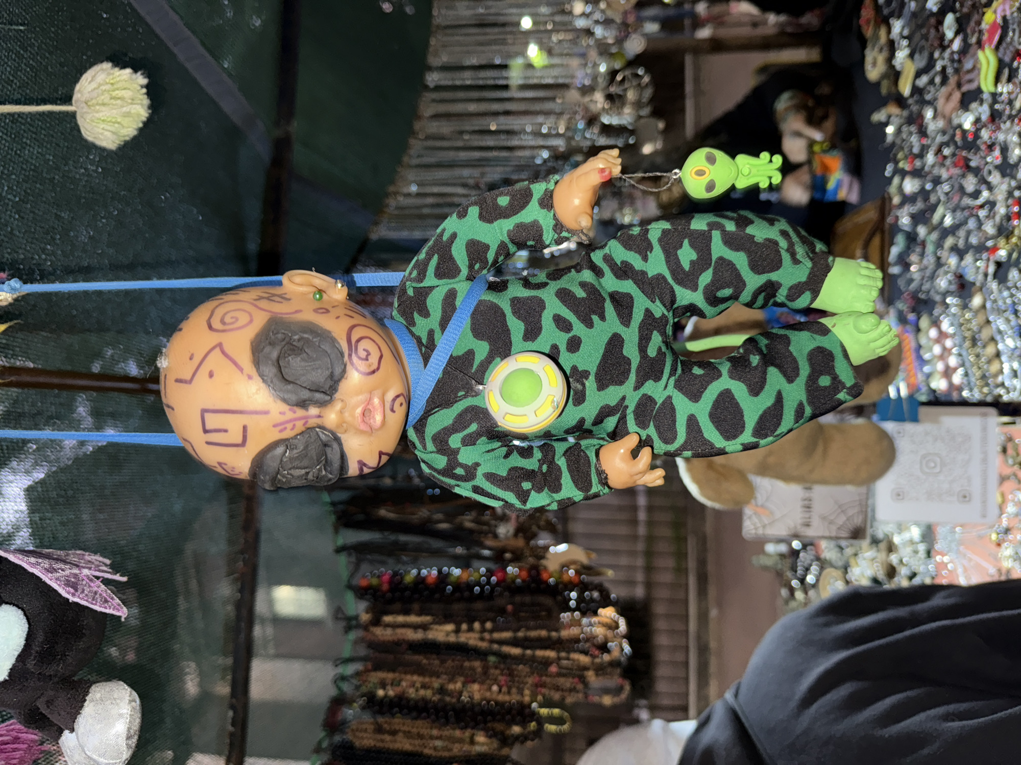 The image shows a doll hanging in what appears to be a street market in Buenos Aires, Argentina. The doll has a tan, round head with black facial markings and small green earrings. It's wearing a green and black patterned jumpsuit and has a blue strap across its chest. The doll's right hand holds a small green alien figurine on a string.
The background is filled with various items commonly found in street markets, including jewelry, beaded necklaces, and possibly other crafts. A person wearing a black hoodie and a white shirt is partially visible in the lower right corner.