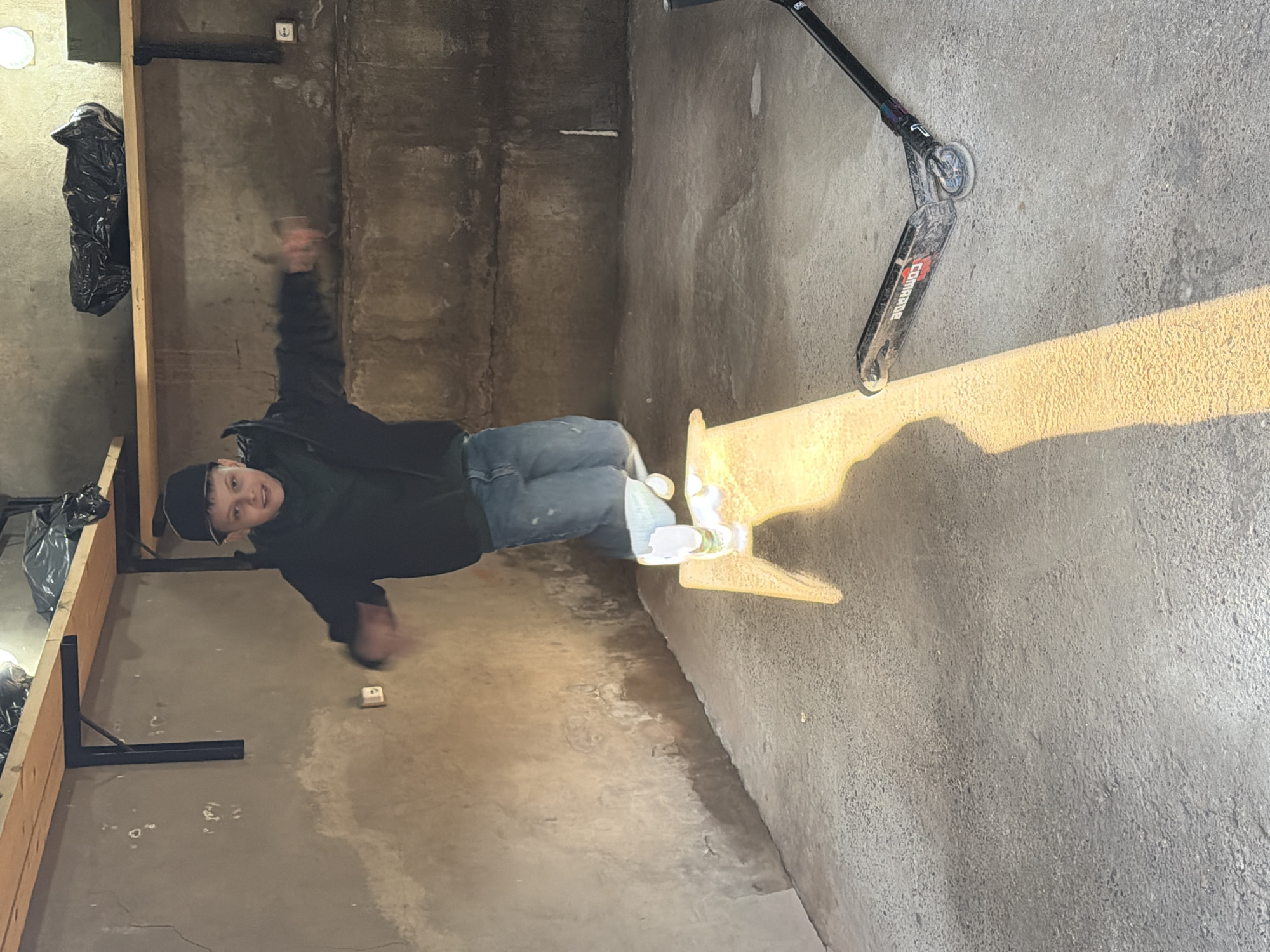The image, rotated 90 degrees clockwise, captures a young person standing on a rough concrete floor within what appears to be a garage, workshop, or similar utilitarian space in Votkinsk, Russia. Strong, golden sunlight streams in from an unseen opening, casting long, sharp shadows across the floor and illuminating the scene, indicating it is daytime.
The central figure, a young individual, wears a dark baseball cap, a dark jacket, and blue jeans, paired with white sneakers. He is smiling broadly at the camera, and his right arm is blurred, suggesting movement or a pose in mid-action.
To the right of the person, a black stunt scooter lies on the concrete floor. The brand name "COMPTRUE" is clearly visible in red and white lettering on its deck. The scooter features black handlebars and wheels.
The background consists of raw, stained concrete walls. Along the left side of the frame, a wooden frame structure runs horizontally, fitted with black metal brackets. A black trash bag hangs from this wooden structure. Higher up on the left wall, a white light fixture is visible, and on the back wall, a white electrical outlet is present. The overall setting conveys an industrial or storage environment.