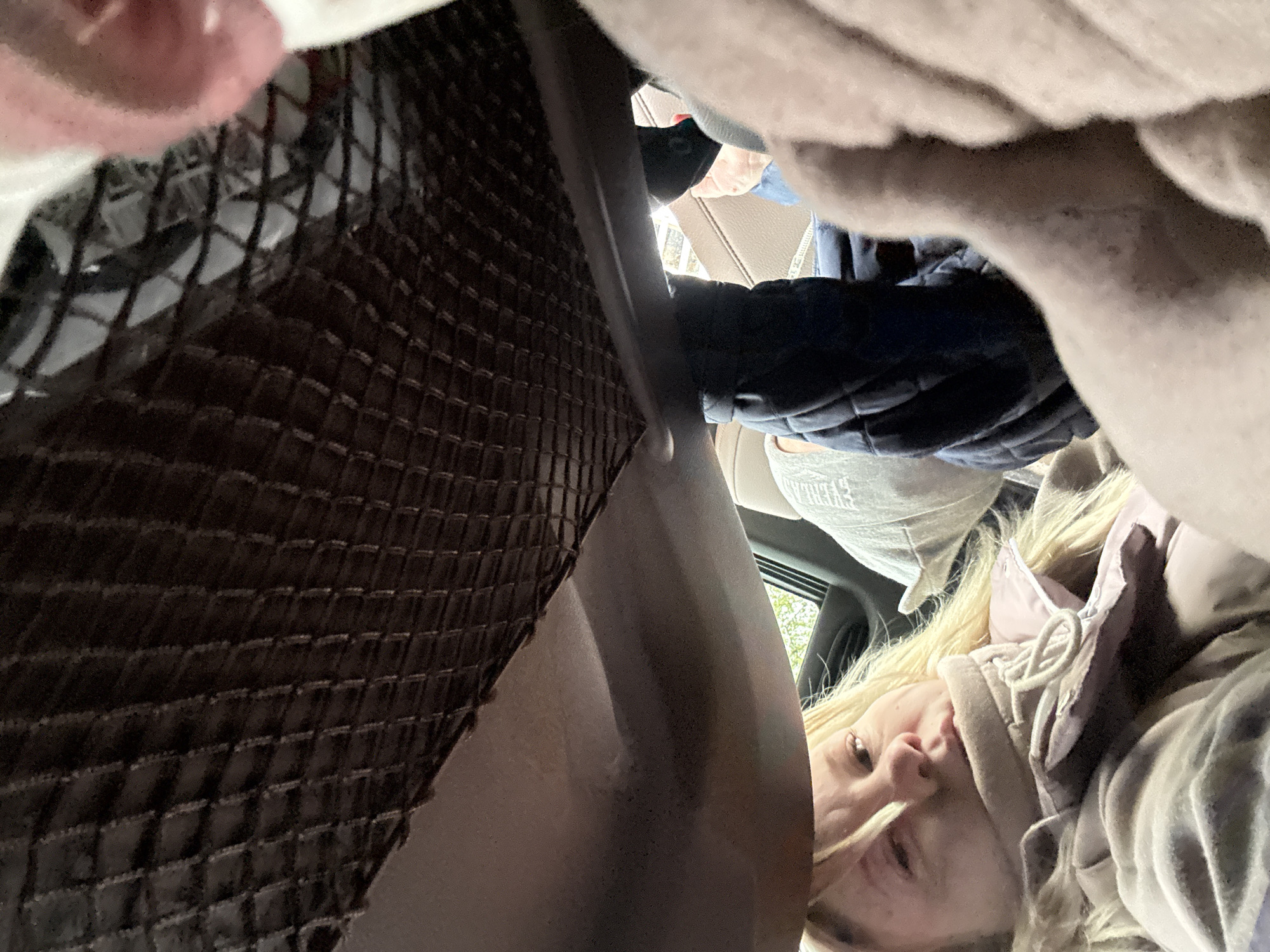 The image shows a view from inside a vehicle, possibly a car, with a person, presumably female, lying back looking upwards towards the camera. She has blonde hair and is wearing a light-colored hoodie. Her face is partially visible. The interior of the vehicle is cluttered with various items, including what appears to be a dark brown woven mat or liner, a dark blue quilted jacket, and a grey hat. The location is not discernible from the image. Without additional context, it is impossible to confirm the location in Chaikovsky, Russia.