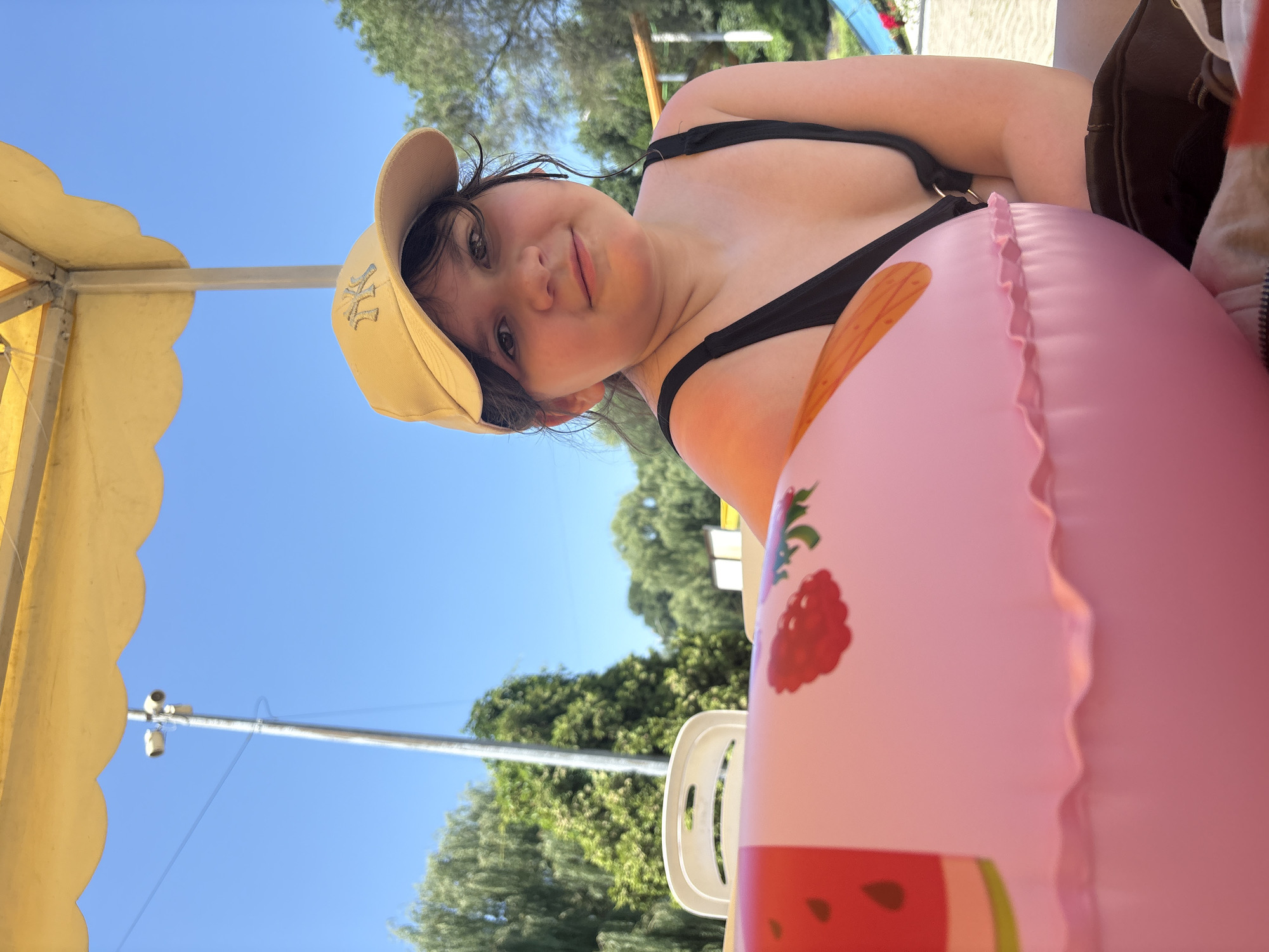 A young girl wearing a black swimsuit and a yellow baseball cap is sitting under a yellow umbrella outdoors. She is looking at the camera with a slight smile. In the foreground, a large pink inflatable ring with fruit patterns is visible. The background shows trees and a clear blue sky. The scene appears to be a sunny day at a recreational area, possibly a park or a beach. There is no identifiable information suggesting the location is in Almaty, Kazakhstan.