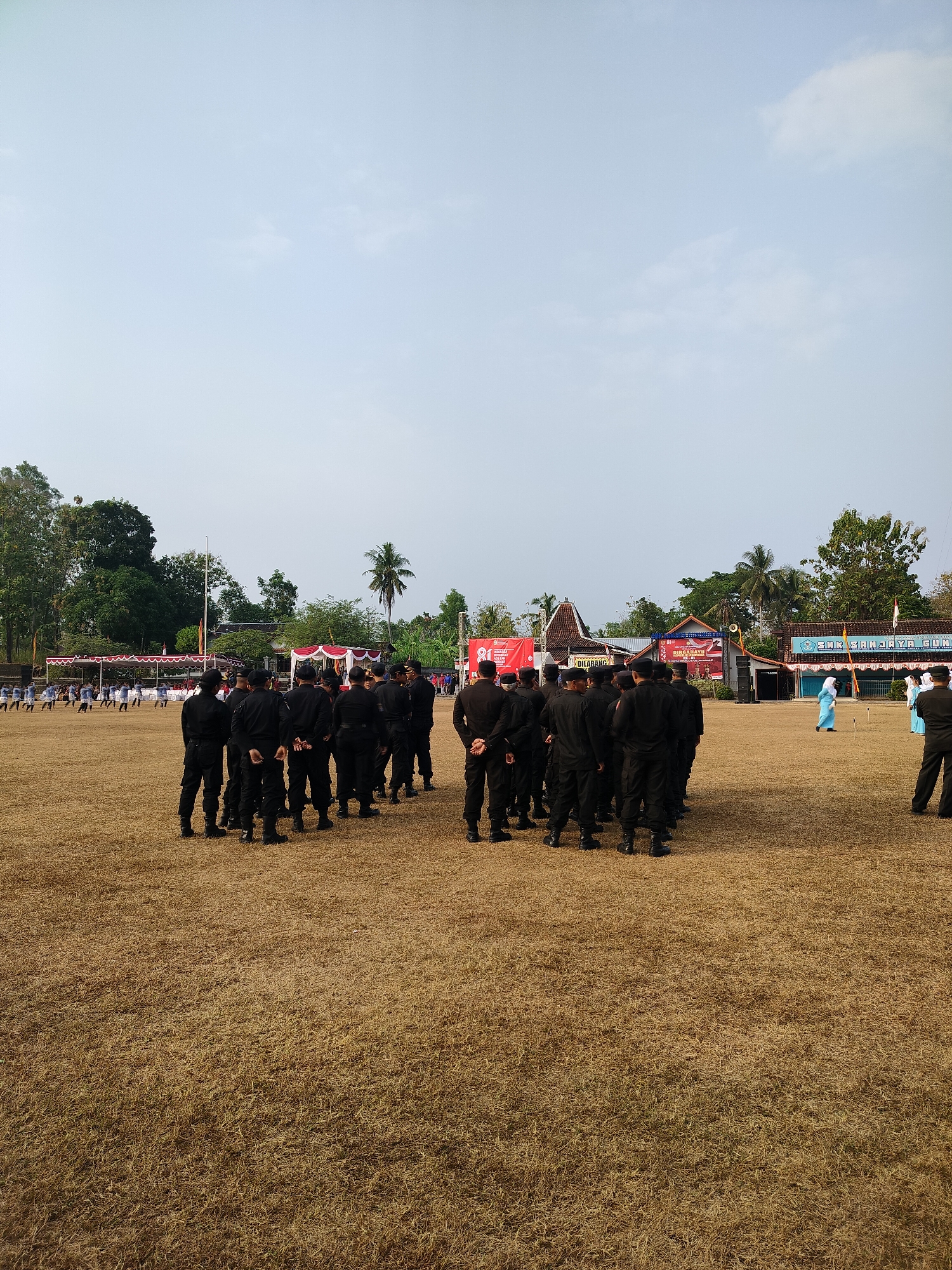 This image depicts an outdoor gathering in Ngawen, Indonesia, likely at a school or public event. A large group of individuals, primarily men, dressed in black uniforms are assembled in formation on a dry, grassy field. They appear to be standing at attention or in a waiting posture. In the background, there are several structures, including buildings with red-tiled roofs, some adorned with banners. One banner prominently displays the number "81" and Indonesian text. Another banner reads "DURGAYU". Further in the background, trees and palm trees are visible against a pale sky with some light cloud cover. Several people in lighter colored attire, including a person in a light blue garment, are positioned further away from the main group. The overall scene suggests an organized event, possibly a ceremony, parade, or training exercise, taking place in a semi-urban or rural setting.