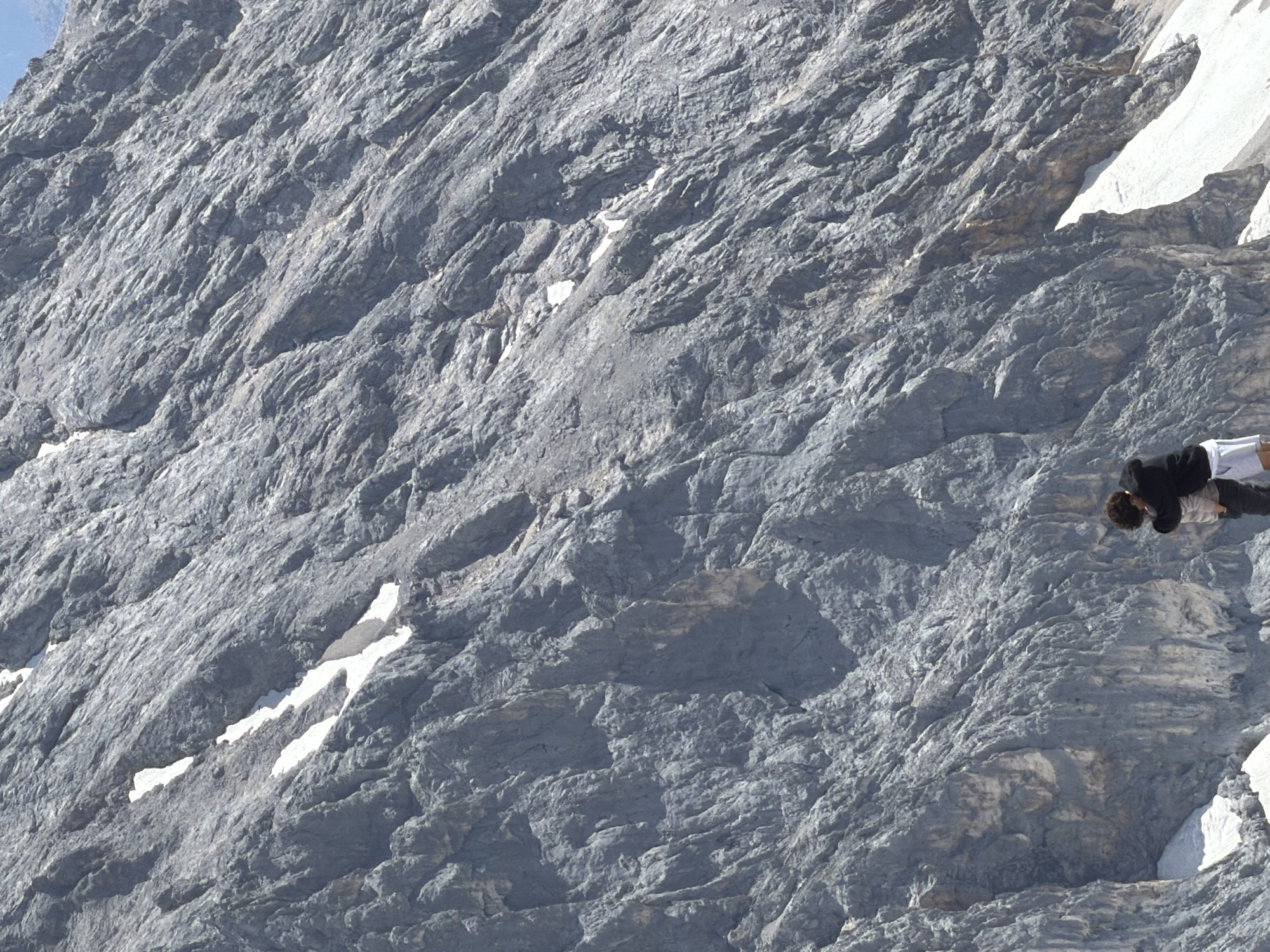 The image shows a steep, rocky mountainside with patches of snow. In the foreground, two people are embracing. The person on the left is wearing a dark jacket and white shorts, and the person on the right is wearing a lighter-colored outfit. The location is the Zugspitze mountain, which is the highest peak in Germany, located near Garmisch-Partenkirchen. The weather appears to be sunny, with a clear blue sky visible at the very top of the image. The overall impression is one of a dramatic natural landscape and a moment of human connection within it.