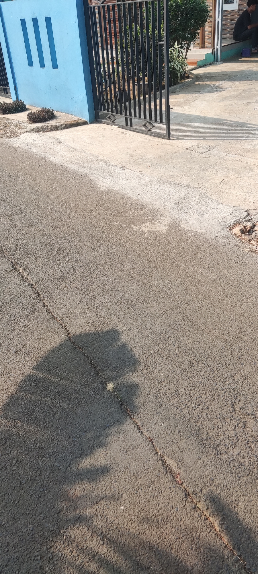 The image depicts a portion of a paved road with a distinct crack running diagonally across it. To the left, a vibrant blue wall with three vertical rectangular cutouts is visible, adjacent to a black metal gate. Small, dark, spiky plants grow in the soil along the base of the wall. The road surface is uneven and appears somewhat weathered. To the right of the gate, a concrete walkway or driveway is present, showing signs of wear and tear, with a section appearing damaged and containing scattered debris. A person is seated on the edge of a slightly elevated platform, partially obscured by shadows and foliage. The overall scene suggests an outdoor environment within a residential or commercial area in Putat, Indonesia.