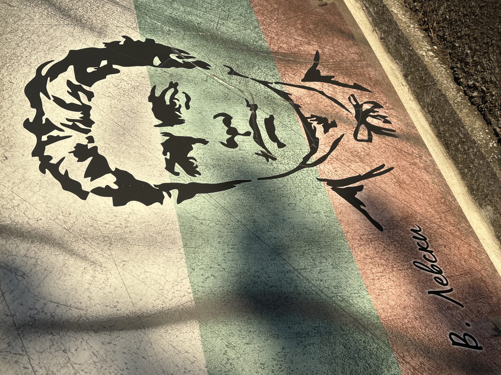 This image shows a stencil art piece on a weathered surface, likely metal or painted concrete, featuring a portrait of Vasil Levski, a Bulgarian national hero. The background is divided into three horizontal stripes, representing the Bulgarian flag: white at the top, green in the middle, and red at the bottom. The portrait is rendered in black silhouette, with Levski depicted with his characteristic hairstyle and attire, including a bow tie. Below the portrait, handwritten in a stylized script, are the Cyrillic letters "В. Левски". The artwork is partially obscured by shadows cast by tree branches. The lower portion of the image shows a concrete curb with some dirt and dried leaves. The scene is located in Enevo, Bulgaria.