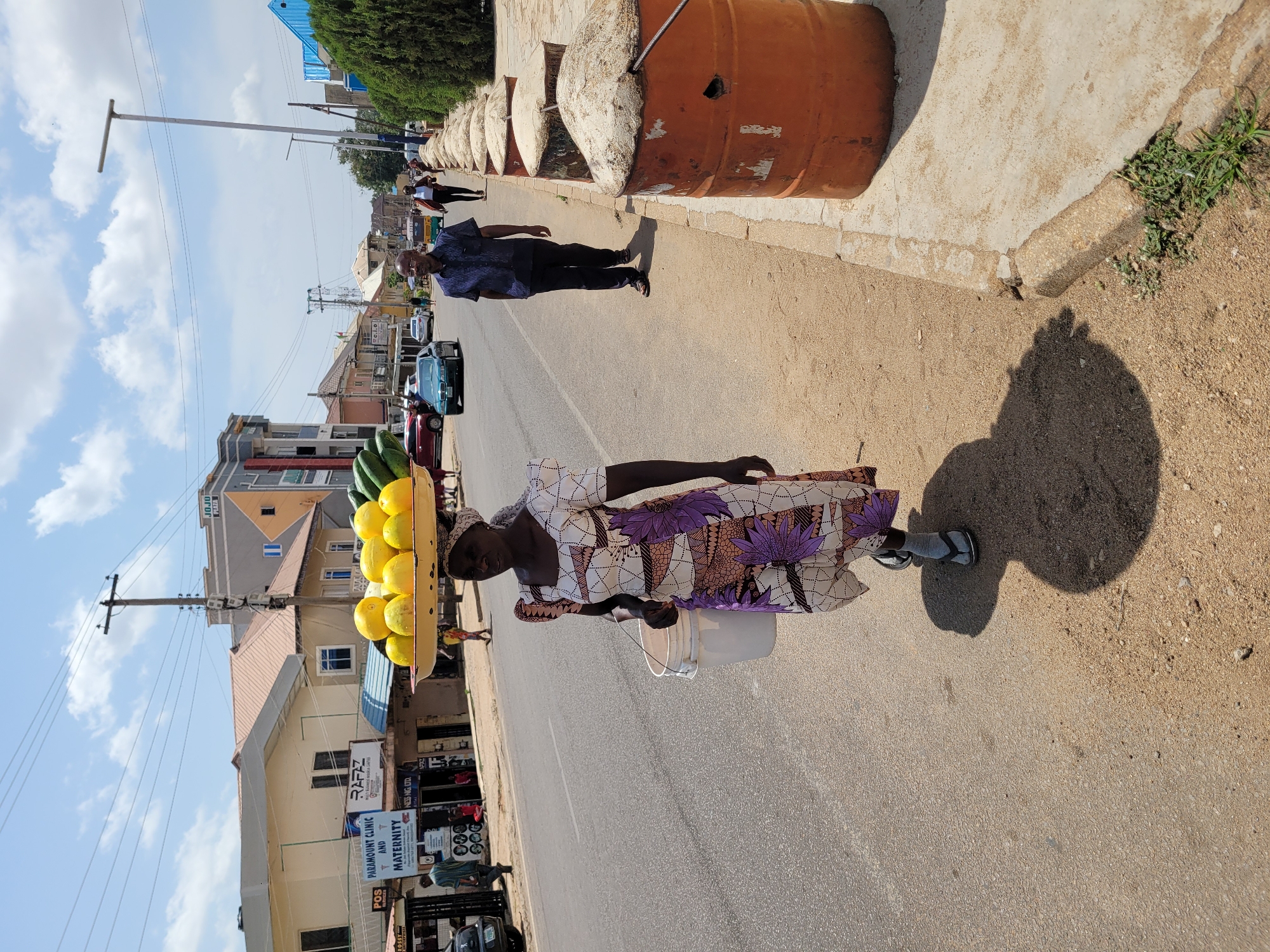 The image shows an older woman carrying a tray of yellow fruits and green vegetables on her head, along a road. She is wearing a patterned dress and sandals, and carrying a white bucket. To her left are storefronts with signs including "PARAMOUNT CLINIC AND MATERNITY," "RAFAZ," and "JOJO PLAZA." Cars are parked along the road. Along the right edge of the image, the ground is sandy with a dark shadow, likely cast by the woman or nearby objects. Behind her, a man in a dark shirt and pants walks along the sidewalk, which is lined with what appear to be orange barrels. Overhead electrical lines are visible. The scene appears to be in the city of Jos, Nigeria, based on the storefront signage.