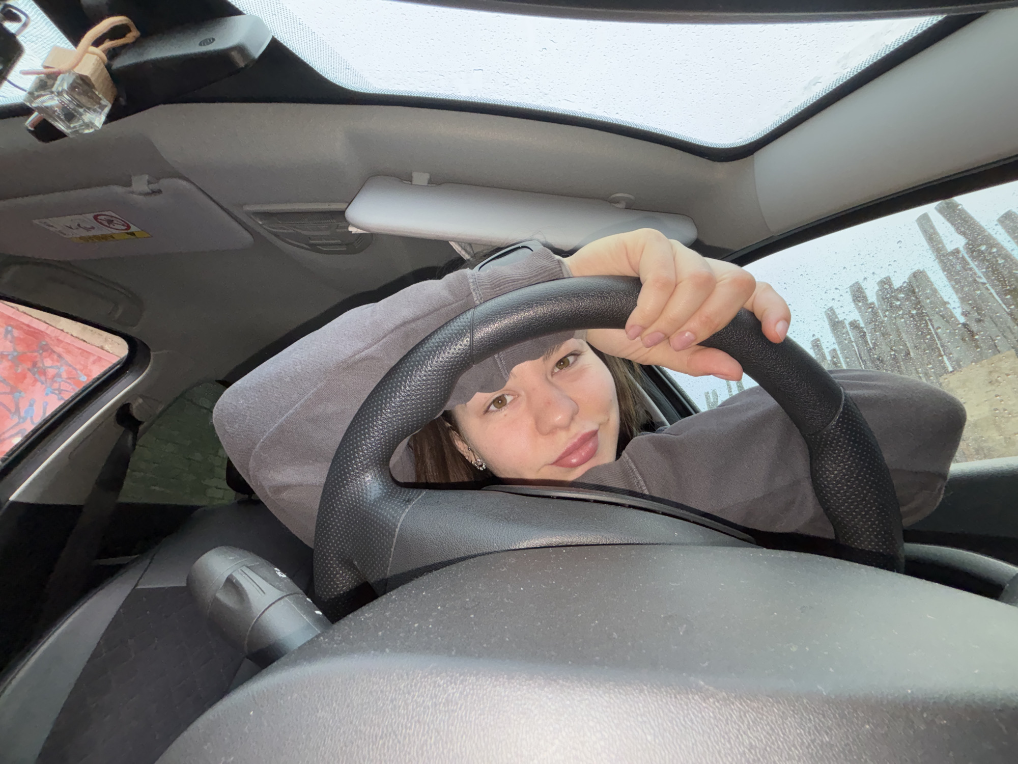 The photograph, presumably a selfie taken inside a vehicle, shows a young woman sitting behind the steering wheel. She is wearing a gray hooded sweatshirt. The car has a sunroof. Outside the car, a rainy or wet scene is visible, with numerous vertical structures, likely posts or pillars, partially obstructing the view. Based on visual analysis and reverse image search, the location is identified as Perm, Russia, specifically near the "Happiness Is Not Far" art installation.