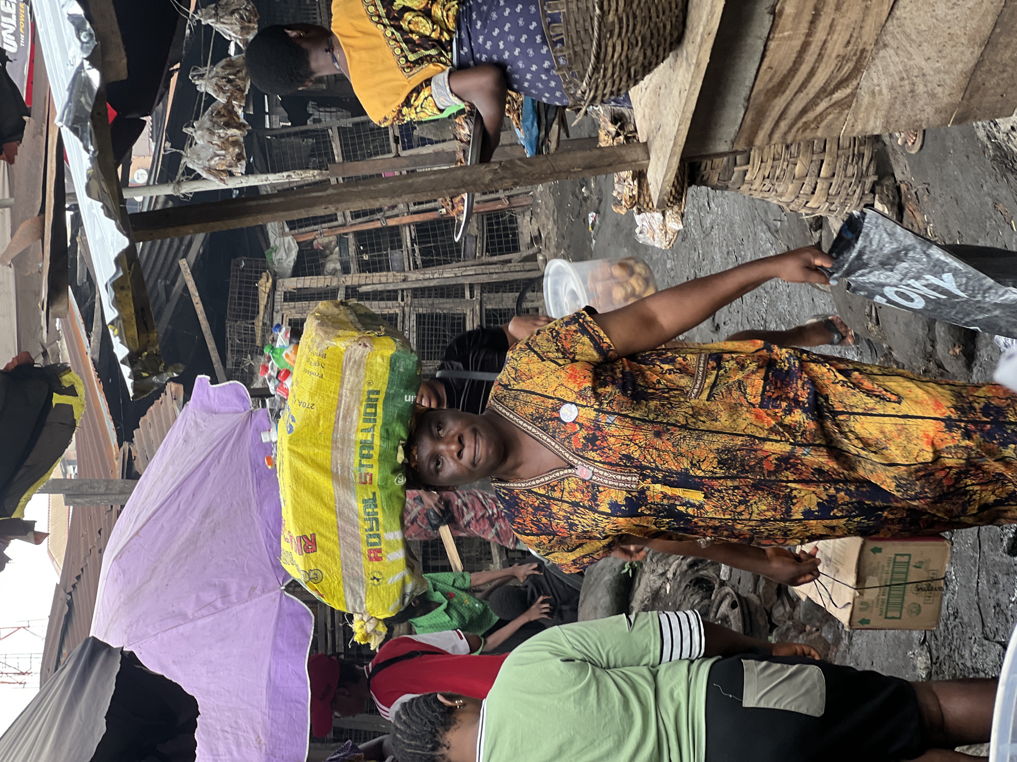 A woman walks through a busy market in Nkpor, Nigeria, carrying a large yellow and green package on her head. She wears a brightly patterned dress. In the foreground, another woman in a green shirt and black shorts walks away from the camera. Behind them, market stalls are visible with various goods, including dried fish hanging overhead and baskets of produce. Other people are seen in the background, with some engaged in transactions. The environment appears to be an outdoor market with corrugated metal roofing and wooden structures. The ground is muddy and cluttered with debris. The overall scene depicts a vibrant and bustling marketplace.
