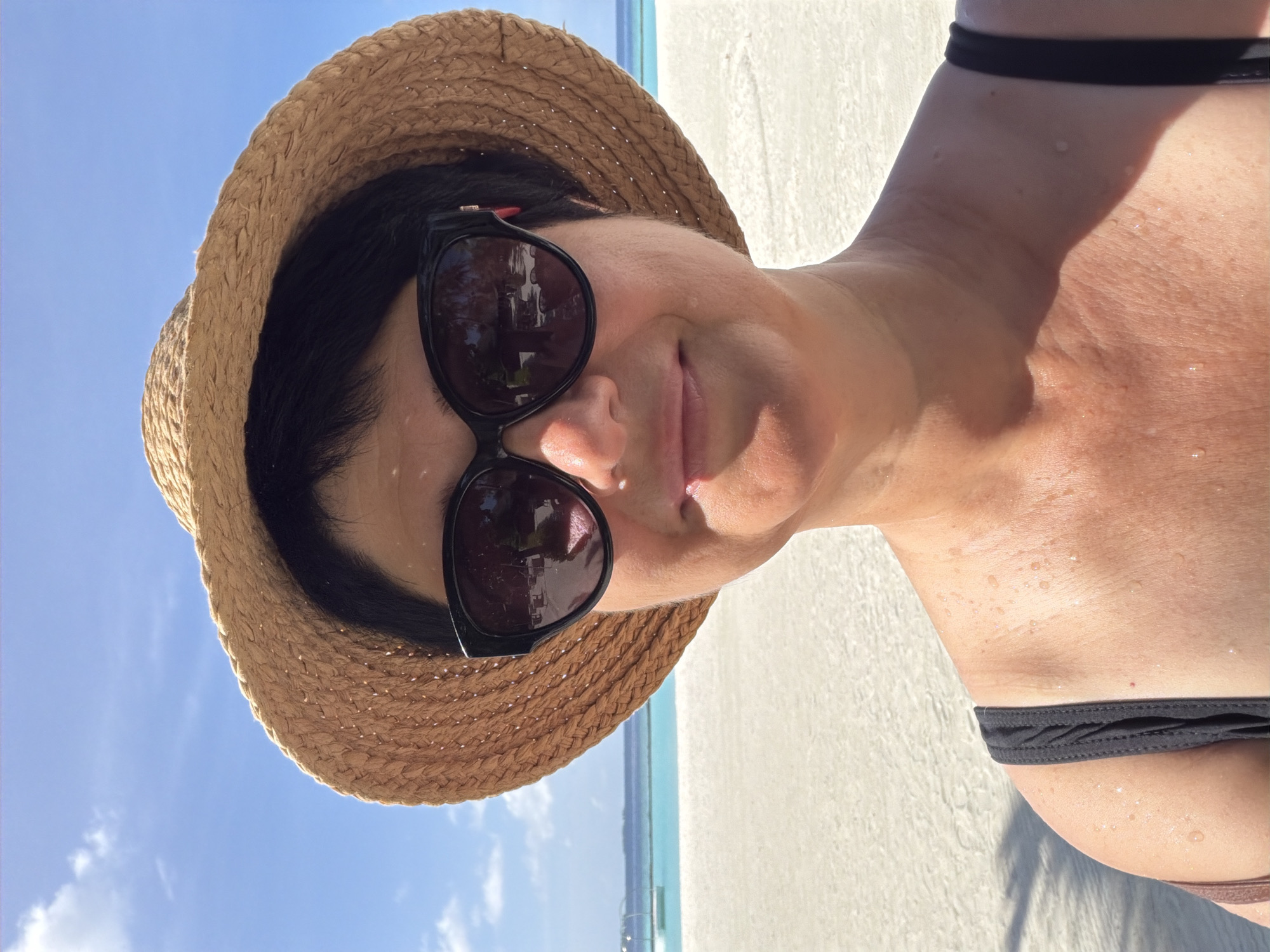 A woman is posing for a selfie on a beach in Halaveli, Maldives. She is wearing a straw hat and sunglasses, and her skin is glistening with water droplets, suggesting she may have recently been in the water. The background shows a white sand beach, turquoise water, and a clear blue sky. There are some structures visible in the distance on the water. The woman's black swimsuit strap is visible.