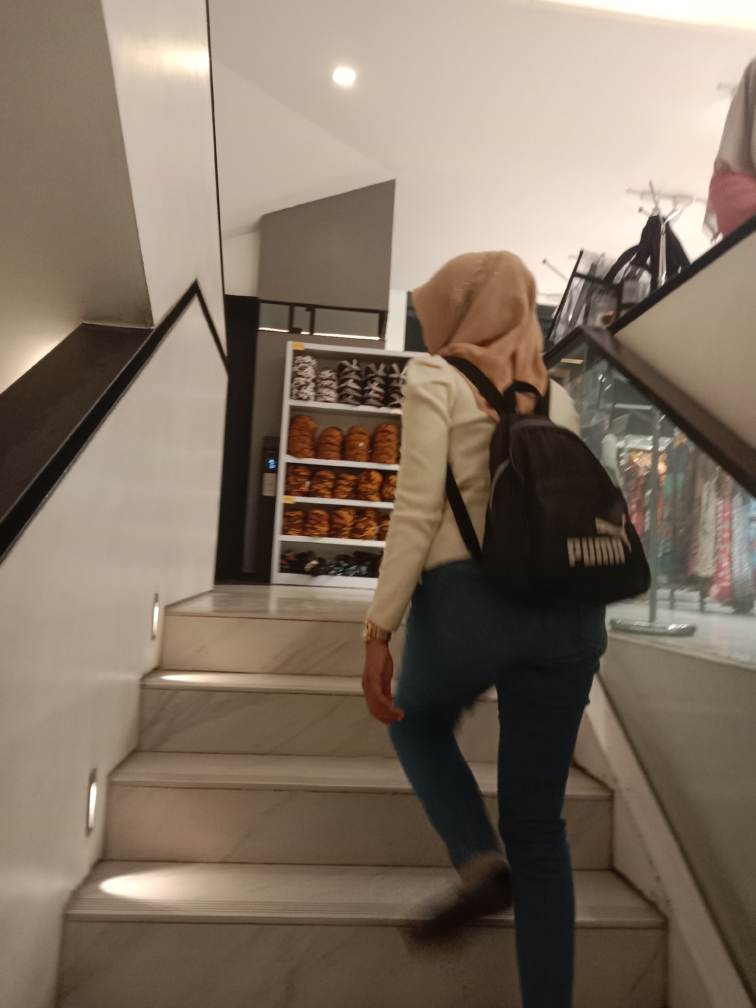 A person wearing a light-colored hijab, a cream-colored long-sleeved shirt, and blue jeans walks up a marble staircase. The person is carrying a black backpack with a white "PUMA" logo. The staircase is illuminated by built-in lights on the left wall. In the background, to the right of the staircase, is a white shelving unit filled with various items, possibly shoes or bags, stacked in rows. Further to the right, a glass partition reveals racks of clothing. The overall environment suggests an indoor commercial space, such as a mall or a store, likely located in Semarang, Indonesia, given the context of the request.