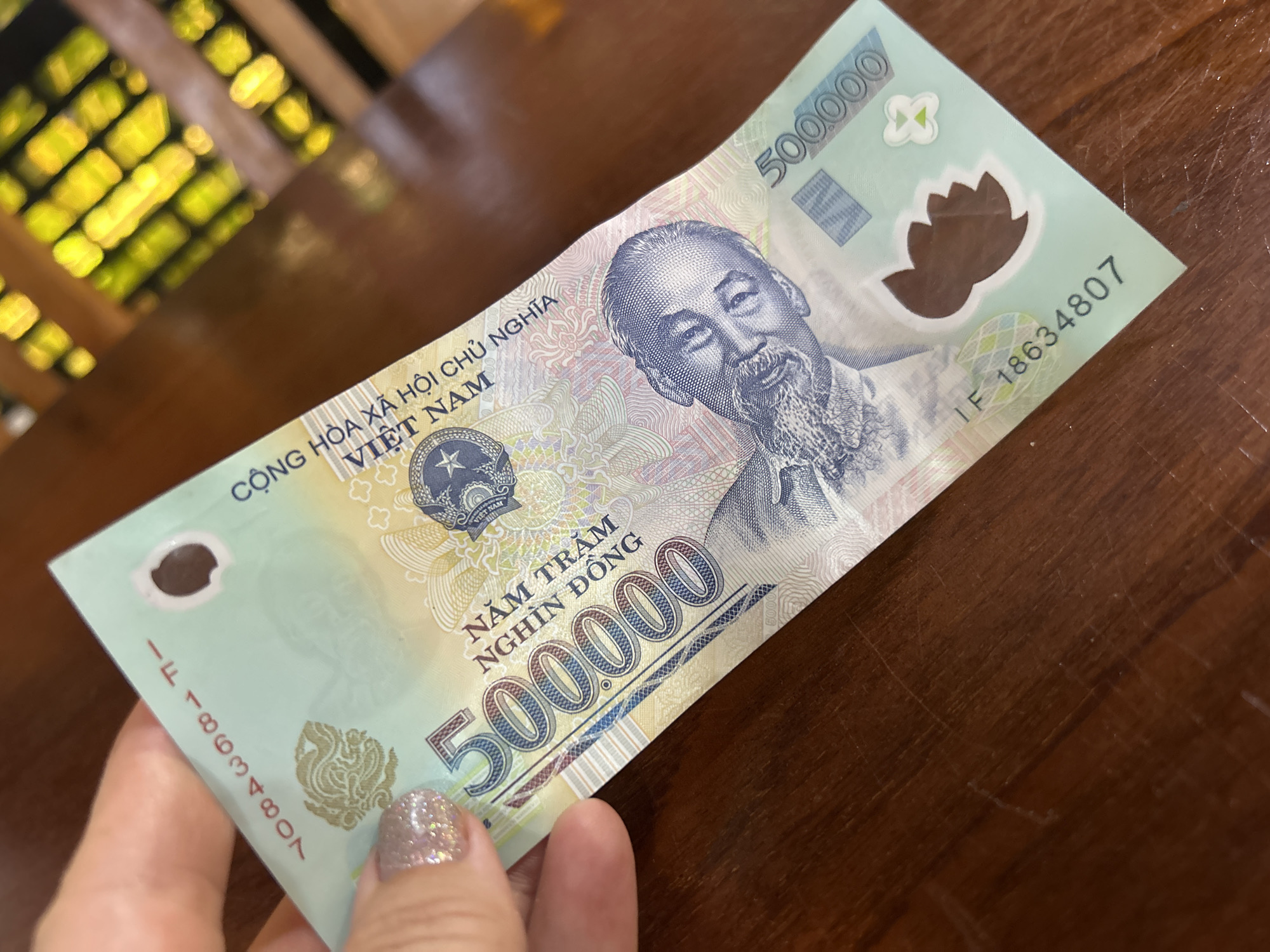 A close-up shot displays a 500,000 Vietnamese Dong banknote resting on a dark brown wooden surface. The banknote is light green and features a portrait of Ho Chi Minh, along with the text "Cộng Hòa Xã Hội Chủ Nghĩa Việt Nam", which translates to "Socialist Republic of Vietnam". The denomination "500.000" is prominently displayed in large numerals. A hand with glittery nail polish holds the edge of the banknote. The background is blurry and shows hints of a brightly lit interior, suggesting it might be a restaurant or cafe, but there is nothing visible to confirm the location in Nha Trang, Vietnam.