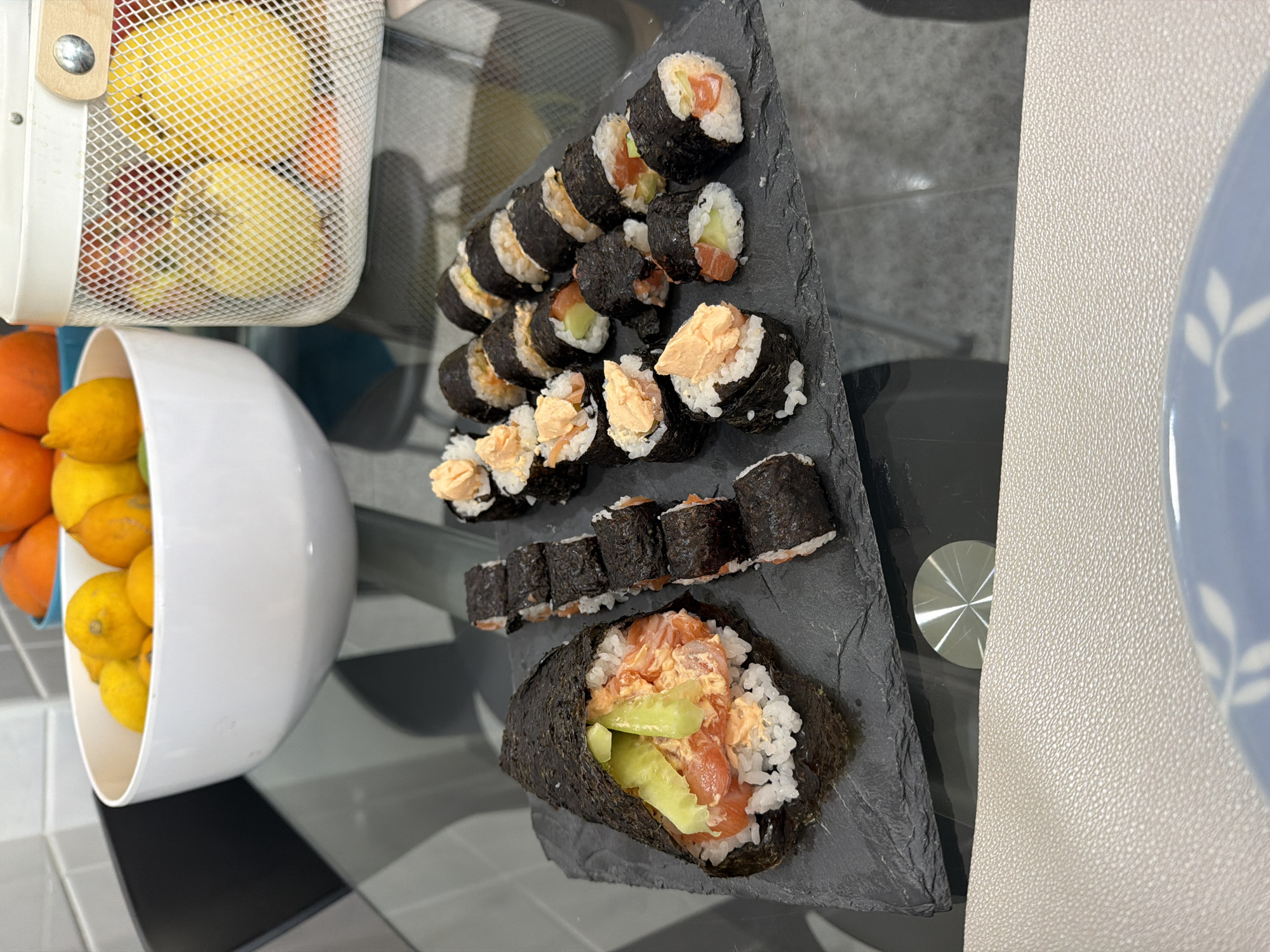 A tabletop in an indoor setting, potentially in Vila Real, Portugal, features a black slate triangular serving platter laden with sushi rolls and a temaki. From left to right, there is a wire basket with yellow citrus fruits, and a white bowl with lemons and oranges. A gray plate, with a white leaf pattern, is partially visible on the right edge of the image. The scene suggests a meal or food preparation setting.