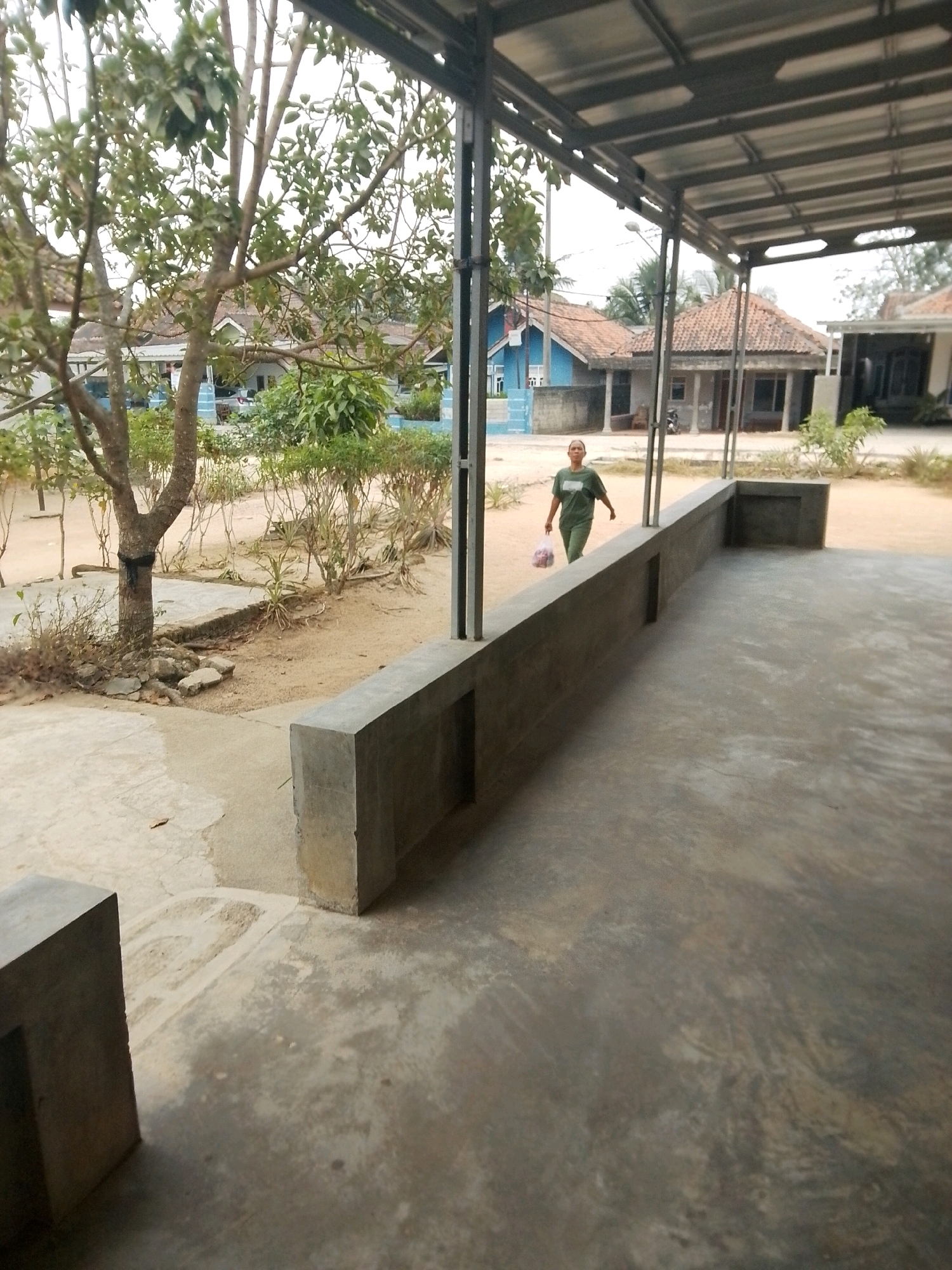 The image depicts a person walking outdoors on a paved area, likely a pathway or courtyard, in front of a building with a covered walkway supported by metal beams. The person, appearing to be an adult female wearing a green t-shirt and pants, is carrying a plastic bag with items inside. Behind the person, several houses with tiled roofs and varying architectural styles are visible, along with vegetation such as trees and bushes. The environment suggests a residential area in Mulyo Haji, Indonesia, characterized by open spaces and simple building structures. The foreground shows a concrete floor and a low concrete wall. The lighting suggests it is daytime.