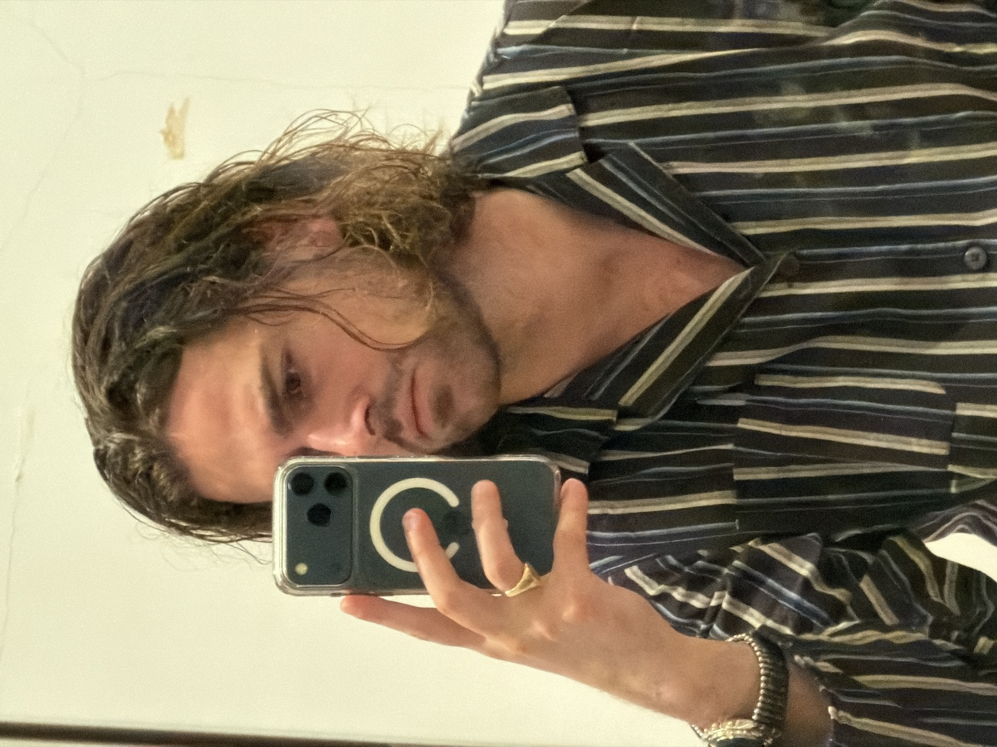 A man with long, wavy, dark brown hair is taking a selfie in a mirror. He is wearing a striped button-down shirt. He is holding a dark blue smartphone with a clear case, and a gold ring is visible on his left ring finger. A silver bracelet is on his left wrist. The background is a plain white wall with a small discoloration mark near the upper left. It's unlikely the location is verifiable as Paris, France based on the provided image details.