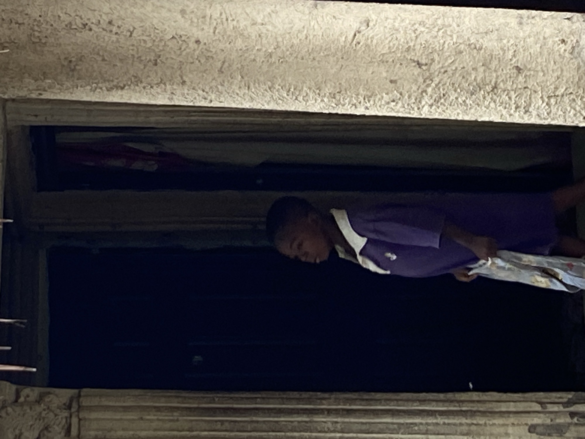 A low-angle shot depicts a person, possibly a child, wearing a dark purple uniform shirt with a white collar and a small emblem, looking down from a window. The child is holding a translucent, patterned object. The window is framed by a concrete structure. The background is dark and indistinct. The location is suggested to be Mararaba, Nigeria.