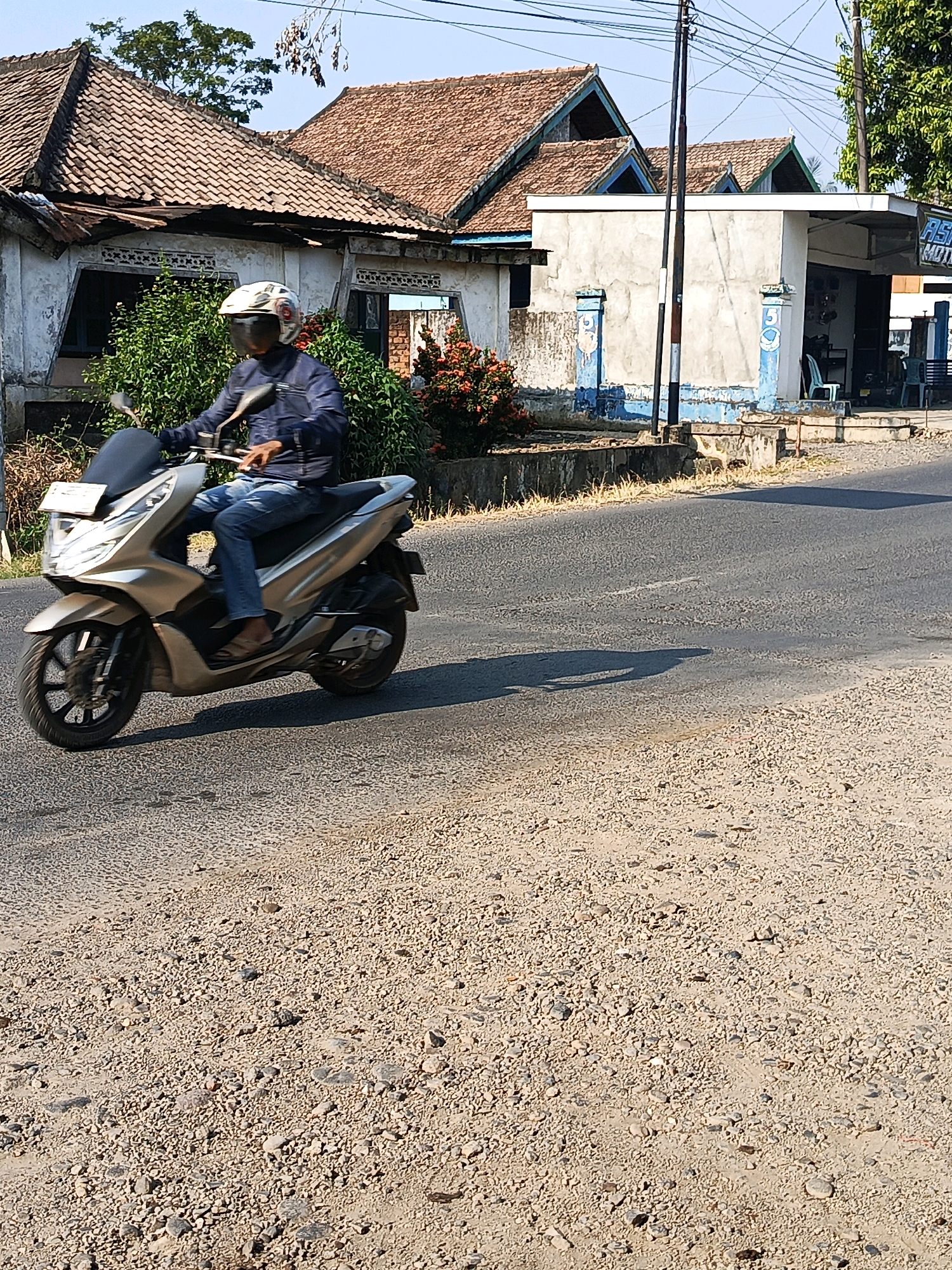 A person is riding a gold scooter down a paved road in the city of Megang Sakti II, Indonesia. The rider, wearing a blue jacket and blue jeans, has a white and black helmet on. In the background, there are houses with red tile roofs and a small building that appears to be a shop. The road is covered in gravel, and there is a shadow cast on the road from the scooter and the rider. The sky is clear and blue.