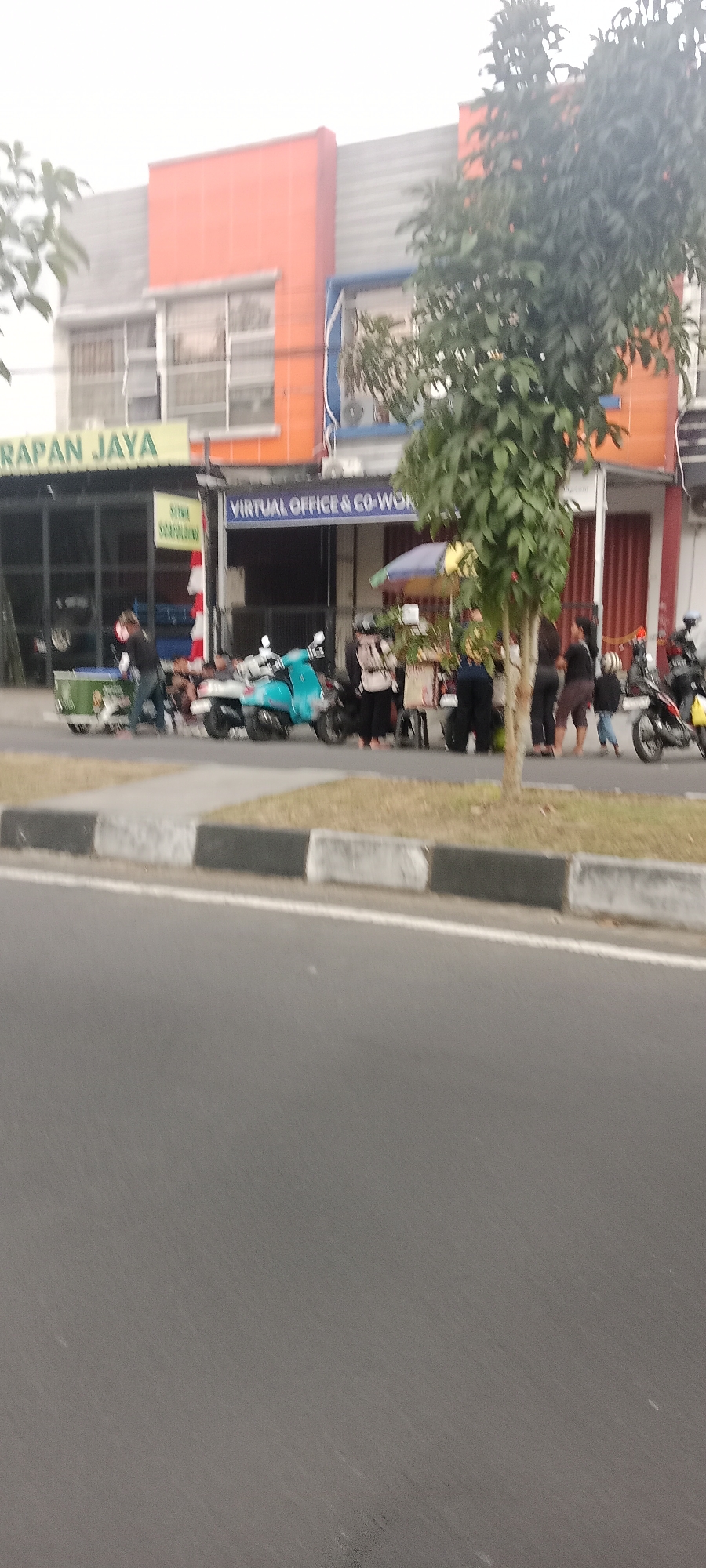 The image depicts a street scene in Ngemplak, Indonesia, featuring a row of businesses along a road. In the foreground, a paved road is visible with a concrete curb separating it from a grassy area. Behind the curb, several people are gathered near a collection of parked motorcycles, including a prominent turquoise scooter. A small food stall with an umbrella is set up, and a few individuals are interacting around it. One person is standing near a green cart. The businesses appear to be a virtual office and co-working space, indicated by a blue sign. Another sign above a black storefront reads "HARAPAN JAYA". The buildings have orange and gray facades. A tree with dense green foliage stands in front of the businesses. The overall impression is a moderately busy commercial area on an overcast day.