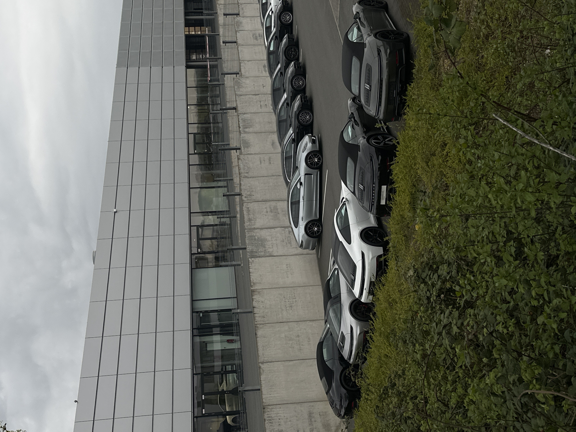 The image shows a row of Porsche cars parked adjacent to a modern building with a gray tiled facade and large windows. The building is likely an office or commercial structure. A dense green hedge borders the cars on one side. The sky is overcast, suggesting a cloudy day. The vehicles appear to be new models, lined up in a parking area. The location is likely Holzwickede, Germany, based on the presence of Porsche vehicles. The photo is taken from a slightly elevated perspective, possibly from a nearby building or structure.