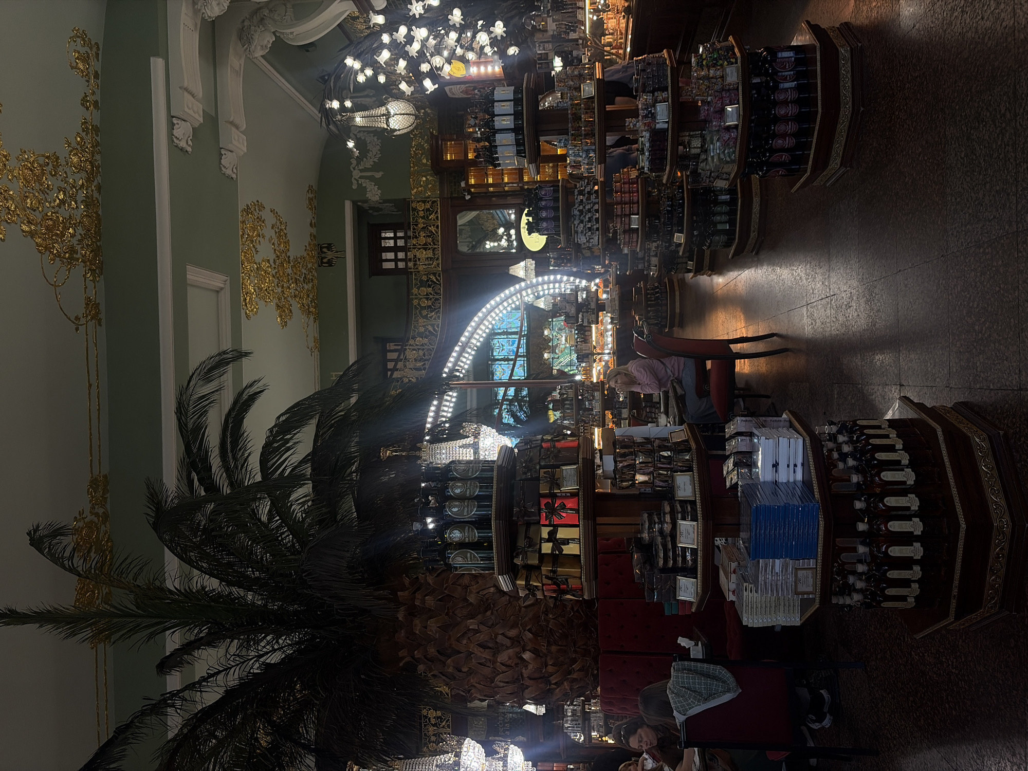 The image depicts the interior of a grand establishment, likely a confectionery or specialty food store, located in Saint Petersburg, Russia. The ornate ceiling features intricate gold detailing and a pale green paint. Dominating the foreground is a large, artificial palm tree with a textured trunk. Surrounding the tree are multi-tiered display shelves laden with various products, including bottles of dark liquid, gift-wrapped boxes, and stacks of books or packaged items.
In the mid-ground, a woman in a pink jacket is seated at a small table. To her left, two other individuals are also seated at a table, their backs to the camera, with one person wearing a patterned scarf. Several other display shelves and tables are visible throughout the establishment, filled with merchandise. The flooring appears to be dark, possibly marble or polished wood, reflecting the ambient lighting.
Several chandeliers, one with a dramatic cascade of light bulbs resembling flowers, hang from the ceiling, contributing to the opulent atmosphere. In the background, tall windows or arched architectural elements with bright, segmented lighting are visible. The overall impression is one of historical grandeur and a well-stocked retail environment. The presence of Cyrillic text on a sign in the background, partially obscured, further confirms the location as Russia.