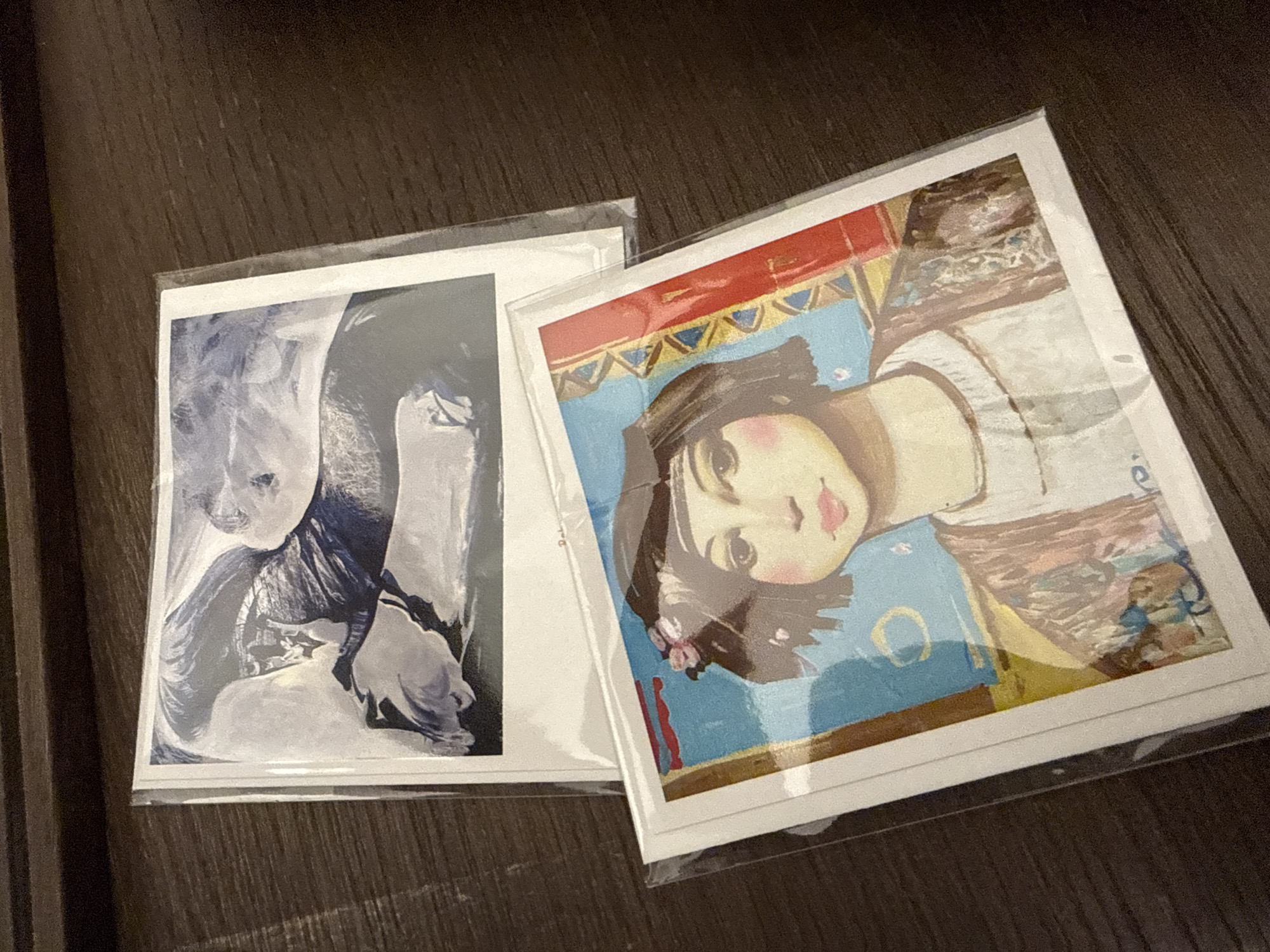 The image shows two framed pictures on a dark wood surface. The picture on the left features a grayscale image of what appears to be two people, possibly embracing. The picture on the right is a colorful portrait of a young woman. Both pictures seem to be wrapped in clear plastic. The location in the city of Krasnoyarsk, Russia is not evident from the image.