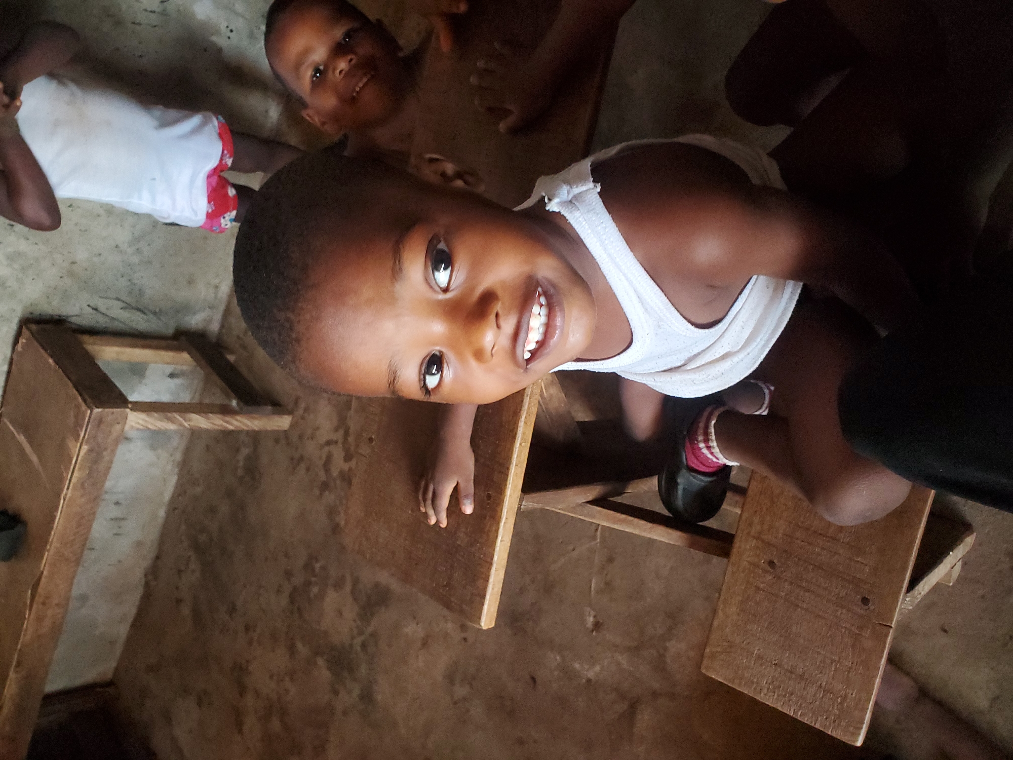 The image shows a young child, likely a boy, wearing a white tank top and smiling at the camera. He is seated on a wooden stool at a simple wooden desk. Another child, also a boy, is visible in the background, also smiling. The setting appears to be a basic classroom or learning environment in Ukwu-Abwa, Nigeria. The room has a worn, possibly concrete floor, and the furniture appears rudimentary. The scene suggests a setting where resources are limited.