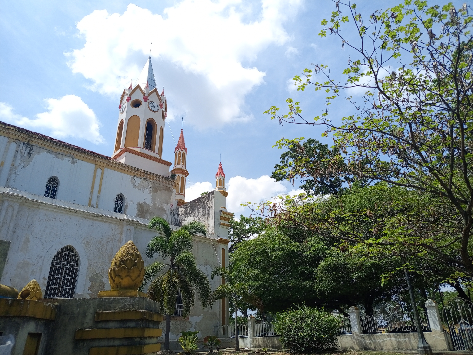 The image depicts a view of the Iglesia Inmaculada Concepción, located in Tinaquillo, Venezuela. The architectural style features a white and yellow facade with a prominent clock tower and multiple smaller towers capped with red conical roofs. A weathered exterior suggests the building is historic. A decorative golden pineapple-shaped structure sits atop a low wall in the foreground. Several trees, including palms, frame the church, with dense foliage creating a natural backdrop. A portion of a wrought-iron fence can be seen near the trees, suggesting a park-like setting. The sky is blue with scattered white clouds.