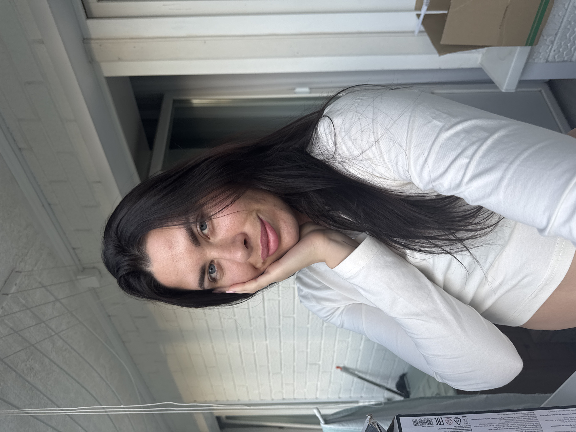 A woman with long, dark hair and blue eyes is pictured in a close-up, outdoor shot. She is wearing a white long-sleeved shirt and resting her cheek on her hand. The background features a white brick wall, a clothesline, and a building with vertical siding. A cardboard box is visible on the right side of the frame. The image appears to be taken during daylight. The location in the city of Chaikovsky, Russia is not discernible from the visual information present in the image.