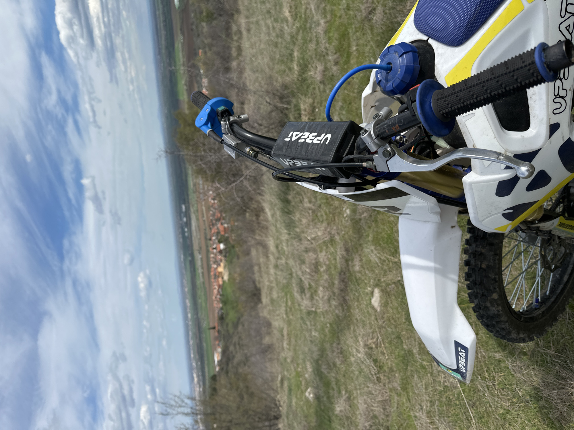 The image shows a portion of a dirt bike, specifically the handlebars and front fender, positioned in a grassy field. The handlebars are black with blue accents, featuring a control module labeled "UPBEAT". The front fender is white and also bears the "UPBEAT" logo. A section of the front tire and spokes are visible.
The background consists of a rural landscape with patches of dry grass and a distant view of a city with visible buildings and sky. The location is not explicitly identifiable within the city of Unknown, Unknown based on the visual information provided.