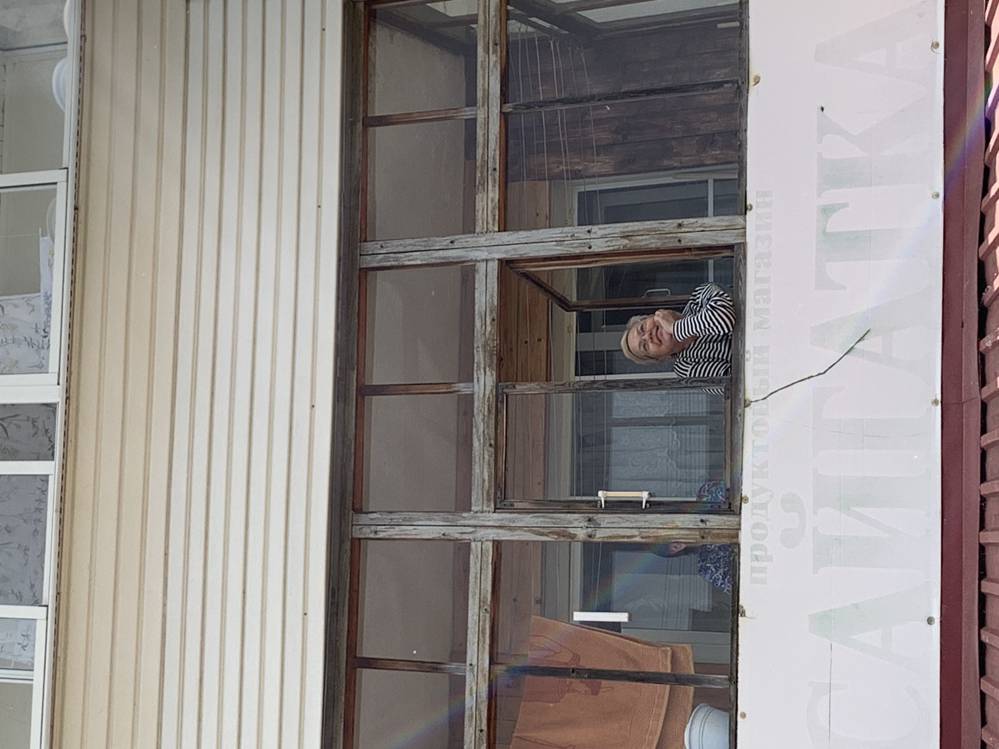 A woman is observed from an exterior perspective leaning out of an open window of a weathered wooden structure, adjacent to a sign vertically displaying text in Russian. The sign is affixed to a building clad in vertically paneled siding. Additional windows are visible within the larger architectural structure. The scene is located in the city of Chaikovsky, Russia.