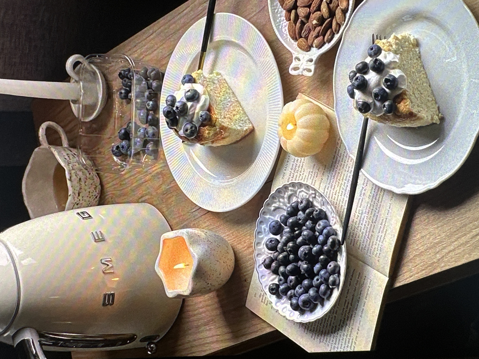 The image depicts a tabletop still life with a focus on desserts and snacks. Two slices of cake topped with whipped cream and blueberries are presented on white plates. A small bowl of blueberries sits adjacent to an open book. A dish of almonds and two lit candles are also visible. A Smeg brand appliance is on the left. A cup is nearby the blueberries. The scene is set on a wooden table. The location is not evident in this image and is not possible to discern from the photograph.