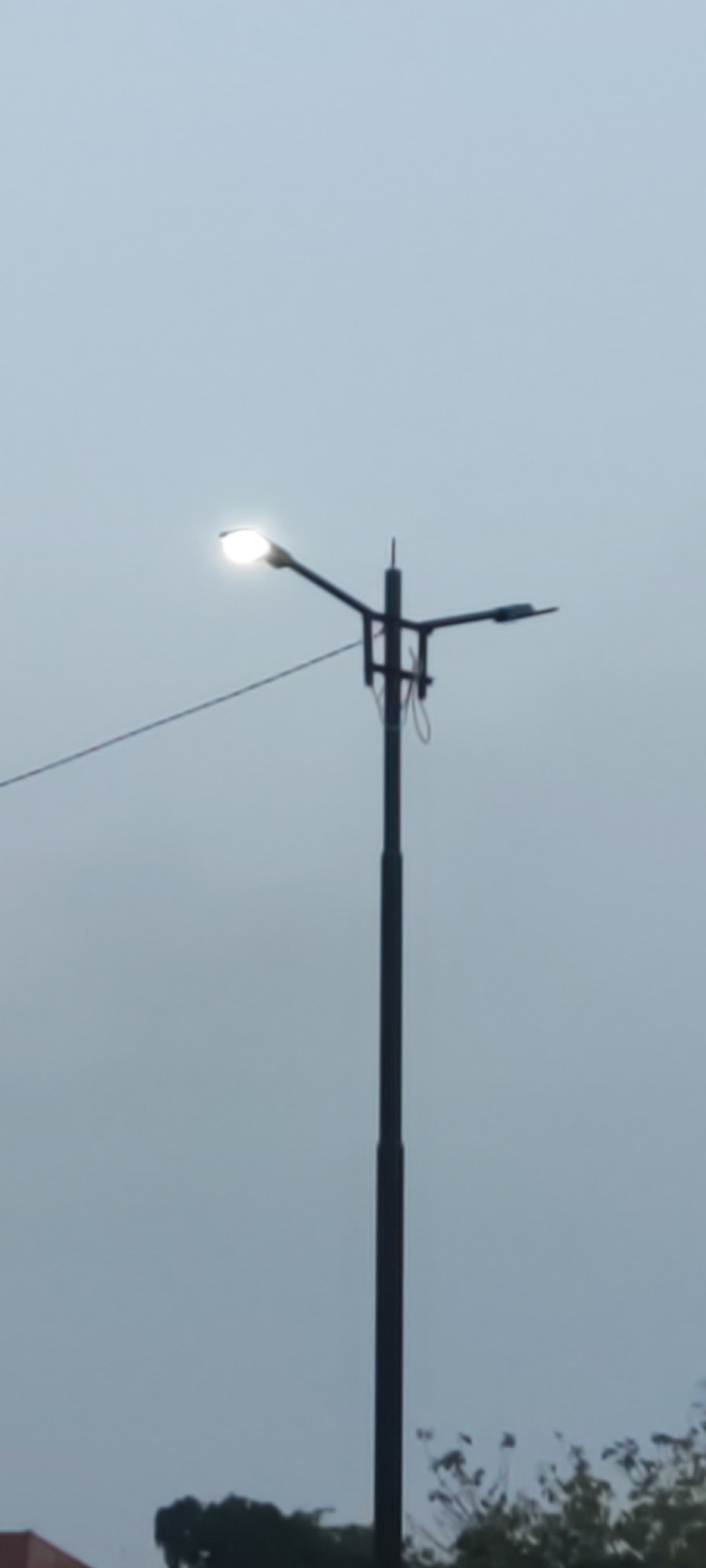 The image shows a street lamp with a lit bulb against a pale, overcast sky. The lamp post is tall and dark, with two arms extending outwards, each holding a lamp. One lamp is brightly illuminated, while the other appears to be off or dimly lit. A wire runs diagonally from the left side of the frame towards the lamp post. In the lower part of the image, the tops of trees and a sliver of a reddish-brown building are visible. The overall scene suggests an outdoor urban or semi-urban environment, possibly during dusk or on a cloudy day. There are no people or animals visible in the frame. The location is identified as Pakusamben, Indonesia.