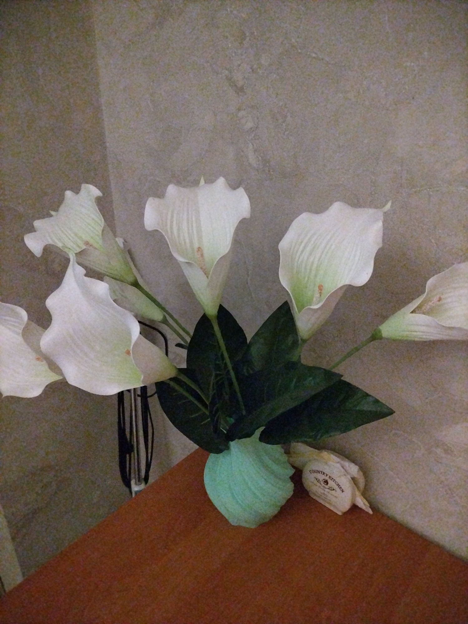 A bouquet of white calla lilies is displayed in a light blue, textured vase. The flowers are arranged with dark green leaves, and the bouquet is placed on a reddish-brown surface, possibly a table or shelf. To the right of the vase, there is a small, light-colored object with text that reads "COUNTRY KITCHEN". The background is a textured, light-colored wall. The scene appears to be an indoor setting. The image quality is moderate, with some graininess visible. There are no people present in the image.