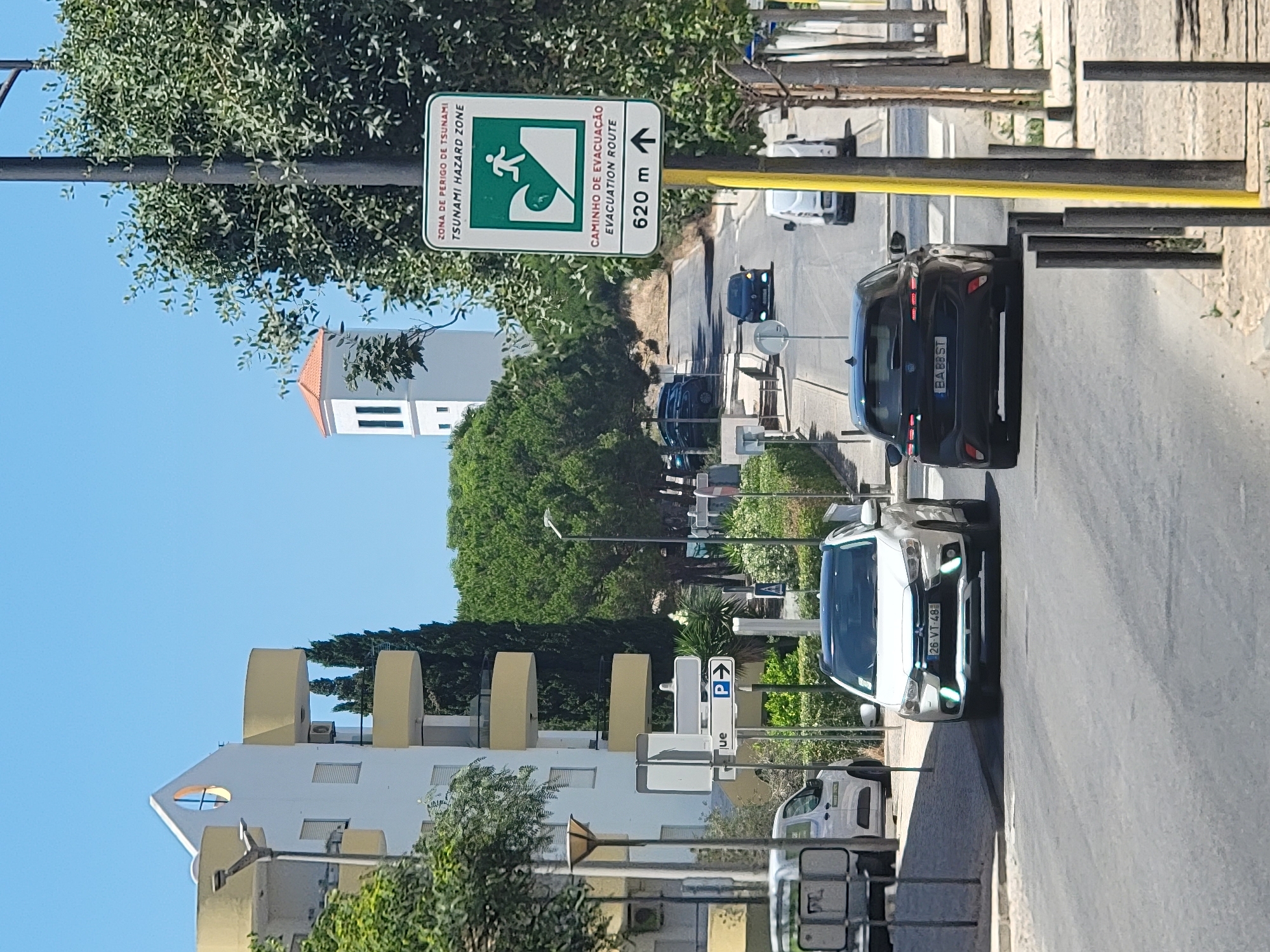 A white Mitsubishi ASX SUV with license plate "26 VT 48" is traveling on an asphalt road in Quarteira, Portugal, on a sunny day. The vehicle is in the foreground, heading away from the viewer. To the right of the SUV, a black Audi is also traveling in the same direction. A green and white sign on a yellow pole indicates a "TSUNAMI HAZARD ZONE" and "EVACUATION ROUTE" 620 meters ahead, with an arrow pointing forward. The road is lined with trees and sidewalk bollards on the right. On the left side of the road, a white building with yellow balconies is visible. A white van is parked on the left side of the road behind the SUV. In the background, a white tower with an orange roof is seen among trees. The sky is clear and blue.