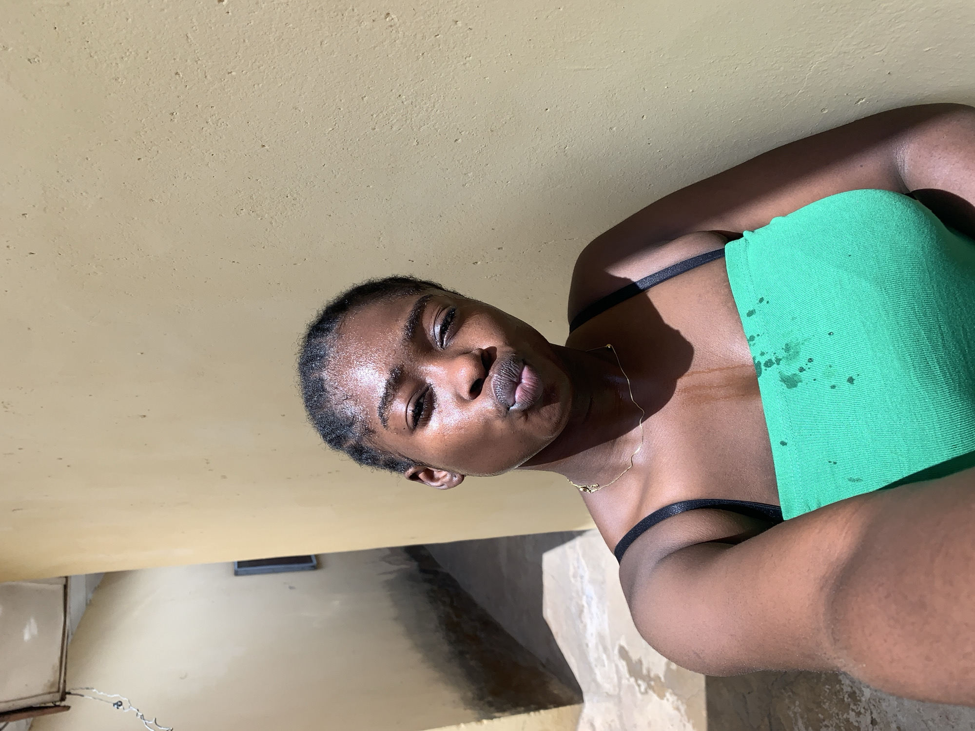 A young woman with dark skin is taking a selfie in a room with a yellow painted wall. She is wearing a green tank top with black straps and a delicate gold necklace. Her hair is braided tightly against her head. The background appears to be a room in a building, with a glimpse of what may be a concrete floor and a metal grate. The location is Rafin Par, Nigeria.