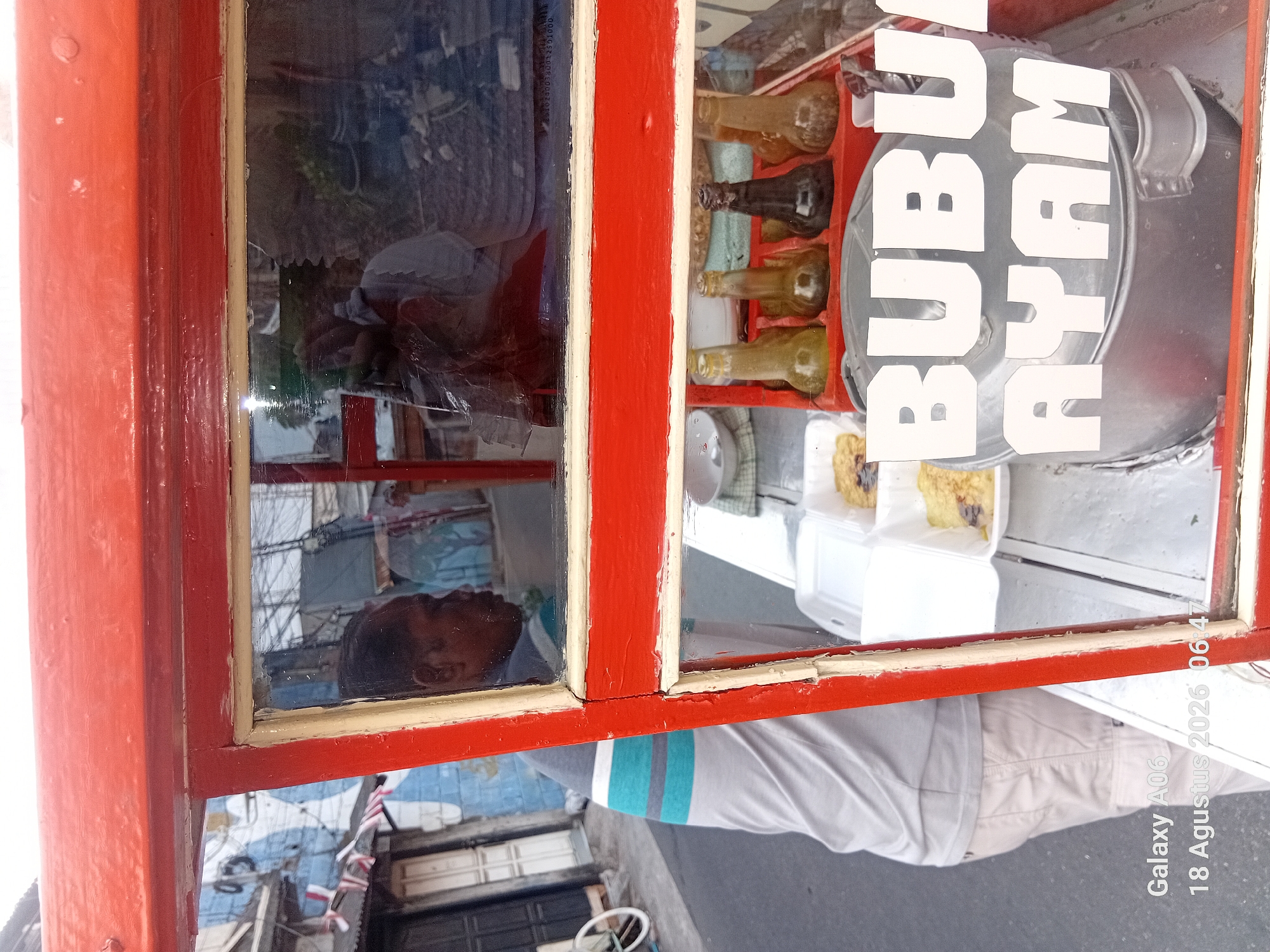 The image captures a close-up view of a red food cart, likely located in South Jakarta, Indonesia, as indicated by the timestamp "18 Agustus 2026". The cart has a glass display window through which various items are visible. Inside the cart, there are several glass bottles containing liquids, suggesting condiments or sauces. Two white styrofoam containers hold food, appearing to be some form of savory dish, possibly with toppings. Prominently displayed on the cart, written in large white letters, is "BUBU AYAM," which translates to "Chicken Porridge," indicating the primary food being sold. In the reflection on the glass, a man can be seen from the shoulders up, wearing a gray t-shirt with blue and gray horizontal stripes. The background reflected in the glass shows a street scene with buildings and what appears to be a mural on a wall, characteristic of urban environments. The overall scene suggests a street-side food vendor operating in an urban setting.
