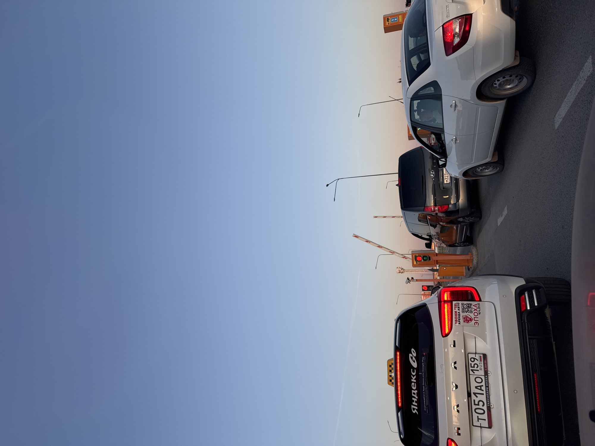 The image captures a scene of vehicles lined up at a toll booth or gate in Bolshoye Savino, Russia. The sky is clear and transitioning in color from a light blue to a subtle orange hue towards the horizon. In the foreground, the rear of a white Haval taxi is visible. It prominently displays the "Yandex Go" logo and the license plate "T051AO 159." Stickers and decals adorn the rear window and trunk, including advertisements for car rentals and taxi services. Behind the taxi, there are other vehicles, including a gray Hyundai with license plate "M323V" and a white Lada Granta. Streetlights and the toll booth structure are situated behind the cars, with traffic lights visible. The barrier arm of the toll booth is in a raised position. The overall setting implies a transportation hub or roadway near Bolshoye Savino.