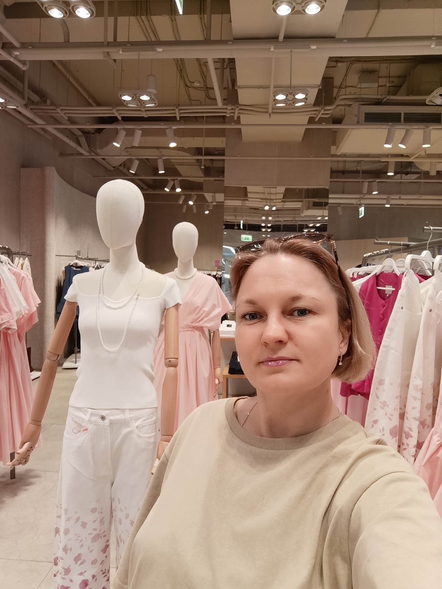 A woman with short blonde hair, wearing a beige t-shirt and sunglasses on her head, is taking a selfie inside a clothing store. Behind her are two mannequins. The first mannequin wears a white top and white pants with floral patterns at the bottom, along with a pearl necklace. The second mannequin is dressed in a light pink dress and a pearl necklace. Racks of clothing in shades of pink and white are also visible in the background. The store has a high ceiling with exposed pipes and numerous lights. The store is in Izhevsk, Russia.