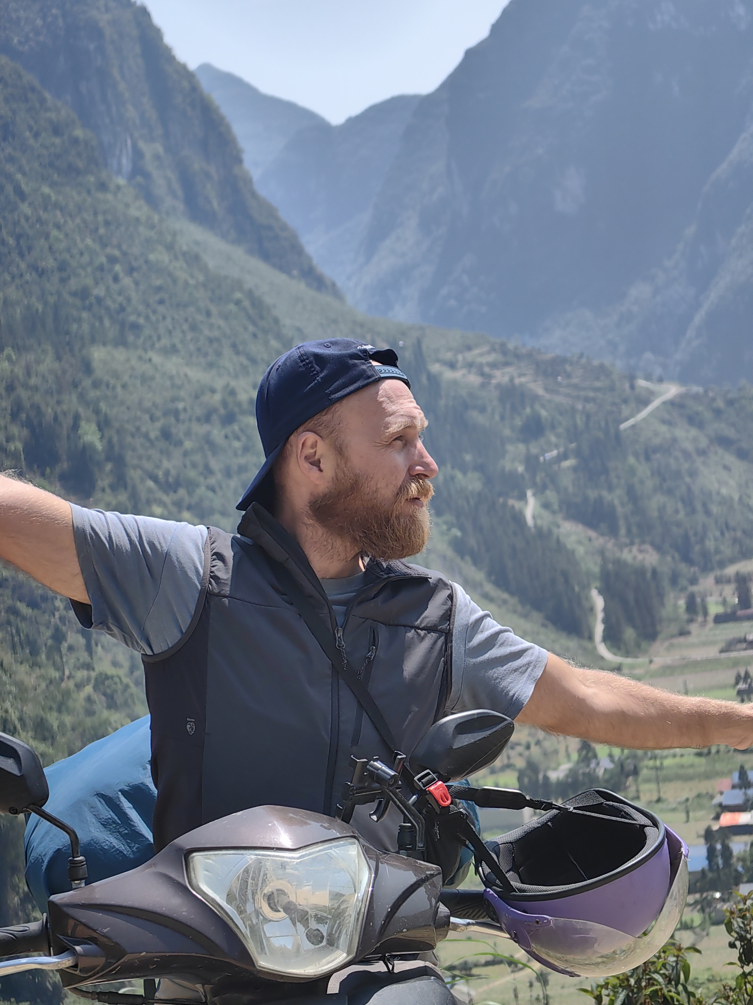A man with a beard and wearing a baseball cap is standing next to a scooter on a mountainside in Phố Bảng Commune, Vietnam. He has his arms outstretched. He's wearing a grey t-shirt and a black vest. A blue bag is strapped to the back of the scooter, and a purple helmet is hanging on the handlebars. Mountains covered in green vegetation form the background.