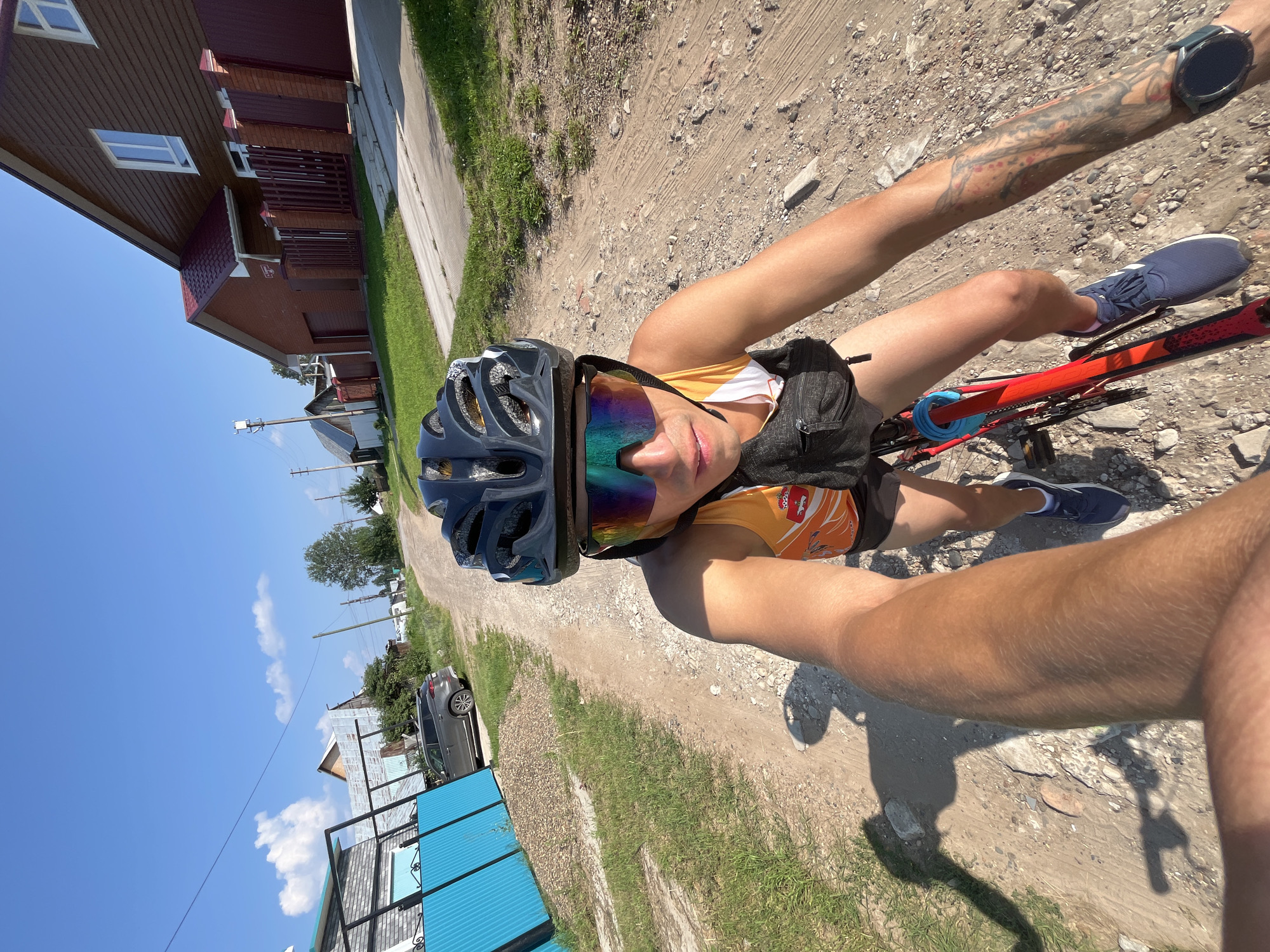 A person is cycling on a dirt road on a sunny day in Chaikovsky, Russia. The person is wearing a helmet, cycling glasses, a yellow and orange athletic top, and dark shorts. They are also wearing a black fanny pack around their waist and blue athletic shoes. A smartwatch is visible on their left wrist. The person's arms and legs are visible, with tattoos on their left arm. The bicycle is red and black.
The scene is a rural or suburban area with houses visible on the left side of the road. Some houses have red roofs and wooden siding. There is grass and trees along the side of the road. The sky is clear and blue with a few white clouds. A car is parked on the side of the road. The road appears to be unpaved and dusty. The time of day appears to be daytime, likely late morning or afternoon given the bright sunlight and shadows. The overall impression is of a recreational cycling activity in a picturesque, sunny environment. No visible text is present in the image.