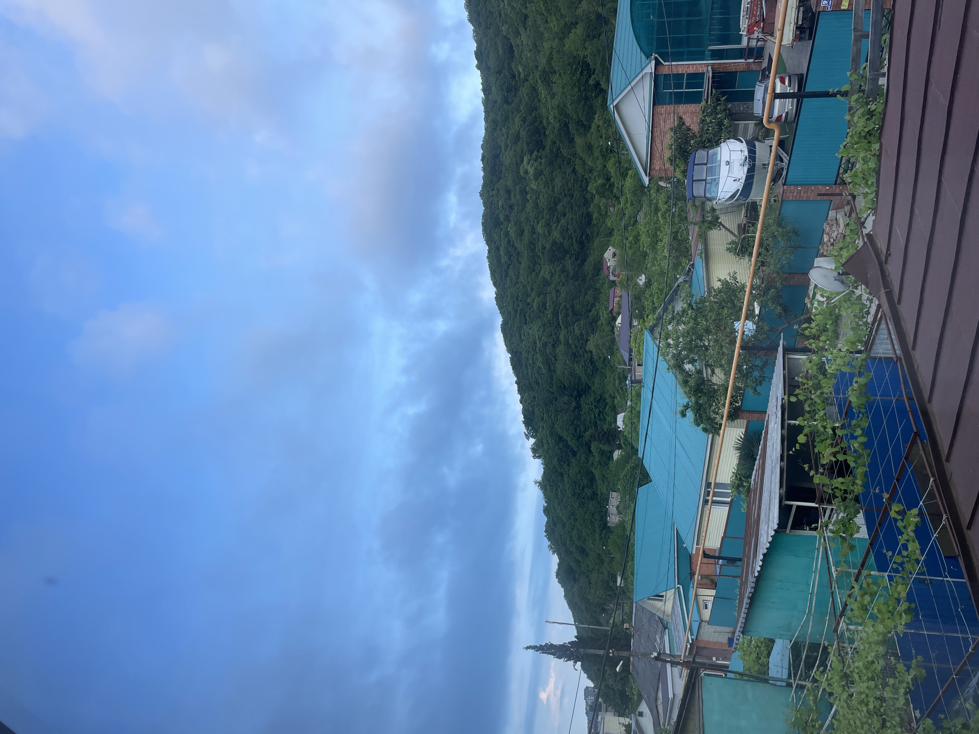 The image captures a view in Olginka, Russia, looking from an elevated position over rooftops towards a lush, forested mountain slope under a cloudy sky. Several buildings with variously colored roofs (blue, brown, green) are visible. On top of one of the roofs, there is a satellite dish and a white boat. The sky occupies a significant portion of the image, filled with clouds.