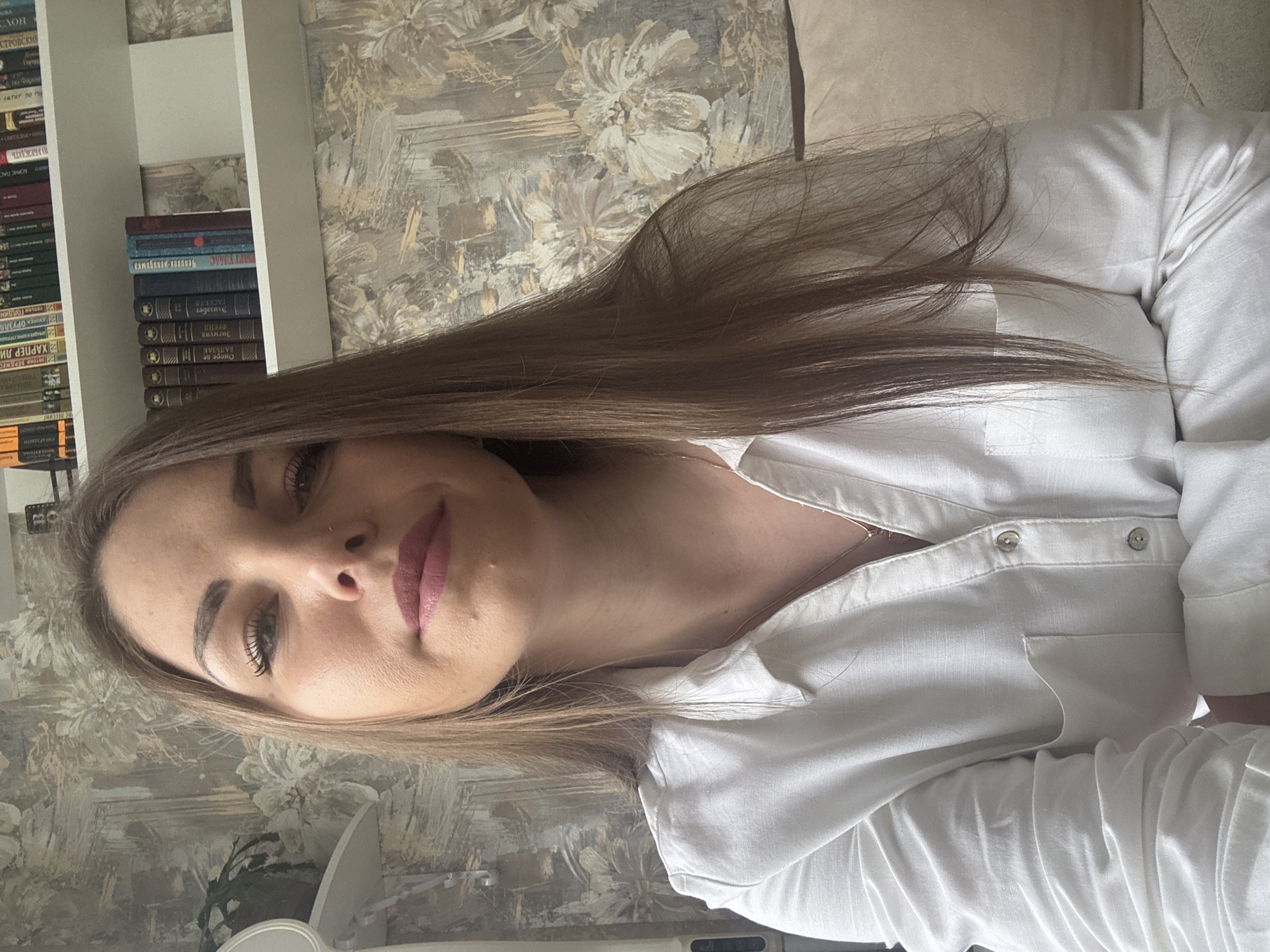 Here's a description of the image:
The photograph shows a woman with long, light brown hair, wearing a white collared shirt with buttons. She is positioned at an angle, with her face tilted towards the camera. She has fair skin, defined eyebrows, visible eyelashes, and pink lipstick.
In the background, there is a bookshelf filled with books. Some book titles are visible in Russian. A wallpaper with a floral pattern is behind the bookshelf. A portion of a beige couch is also visible.
It's not possible to verify the exact location in the city of Раздолье, Russia, based on the visual information in the image alone.