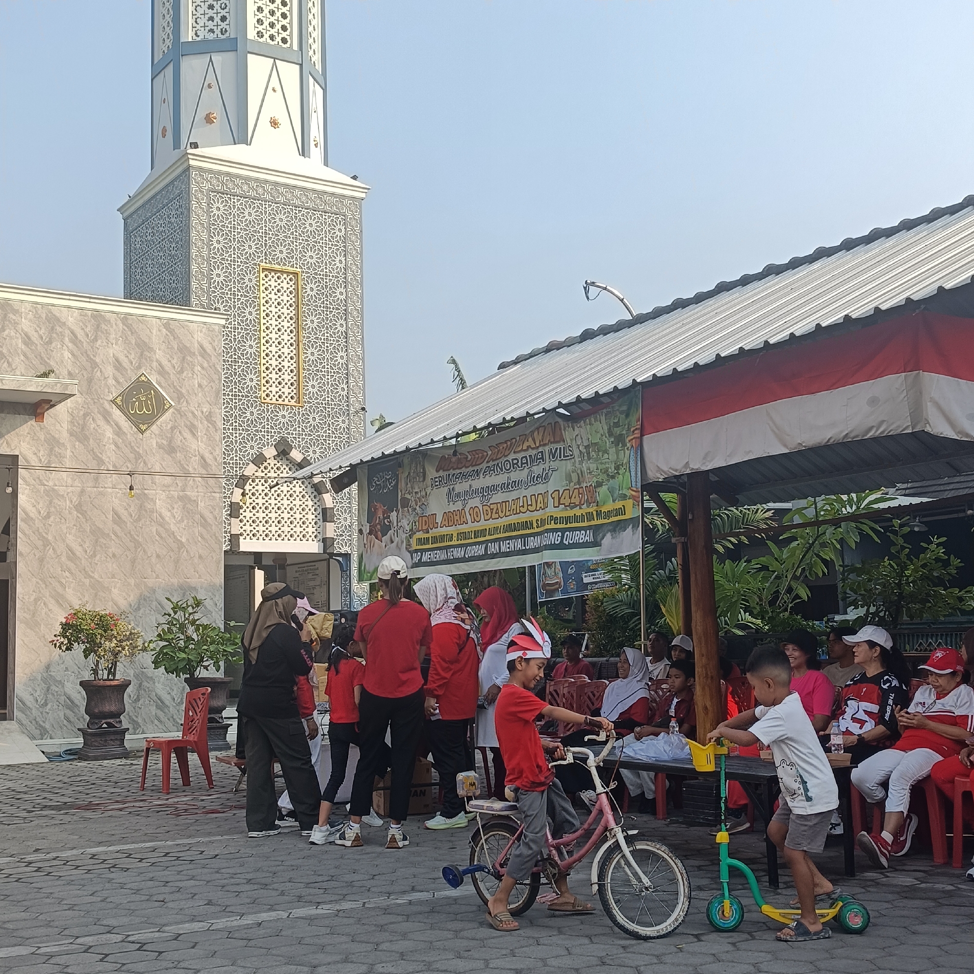 In Madiun, Indonesia, a group of people, predominantly women and children, are gathered outdoors in front of a mosque and a covered area with a red and white striped awning. The mosque features a tall, intricately patterned minaret and a facade with Arabic calligraphy. A banner hangs on the covered area, advertising an Eid al-Adha event, mentioning "Masjid Panorama Mils" and "9 Dzulhijjah 144H". Several children are engaged in activities: one boy is riding a pink bicycle with training wheels, another is on a green and yellow scooter, and others are standing or sitting. Adults are seen observing, some are dressed in red shirts. The environment consists of a paved courtyard with some potted plants and red plastic chairs. The overall scene depicts a community gathering, likely for a religious observance.