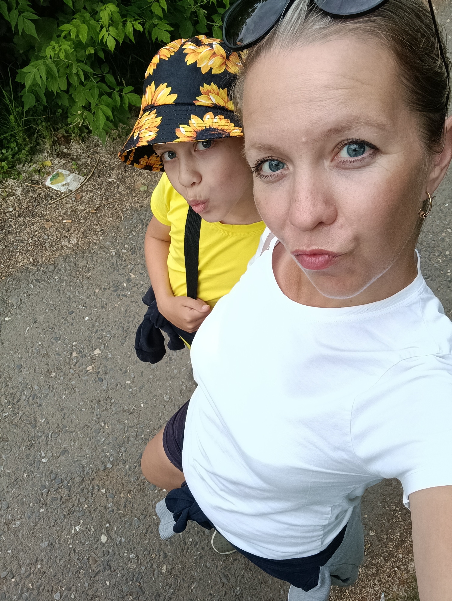 A selfie taken from a high angle shows two people, an adult and a child, standing on a paved surface with a wooded area in the background. The adult, in the foreground, is wearing a white t-shirt and dark shorts. They have blonde hair pulled back and are wearing sunglasses on their head. The child, slightly behind and to the left, is wearing a yellow t-shirt and a bucket hat with a sunflower pattern. The child is also wearing a black strap across their chest. Both individuals have their mouths puckered. The ground appears to be asphalt mixed with gravel, and there is some green foliage and a discarded wrapper visible to the left of the frame. The scene suggests an outdoor setting, possibly a park or a pathway. The location is the city of Chaikovsky, Russia.