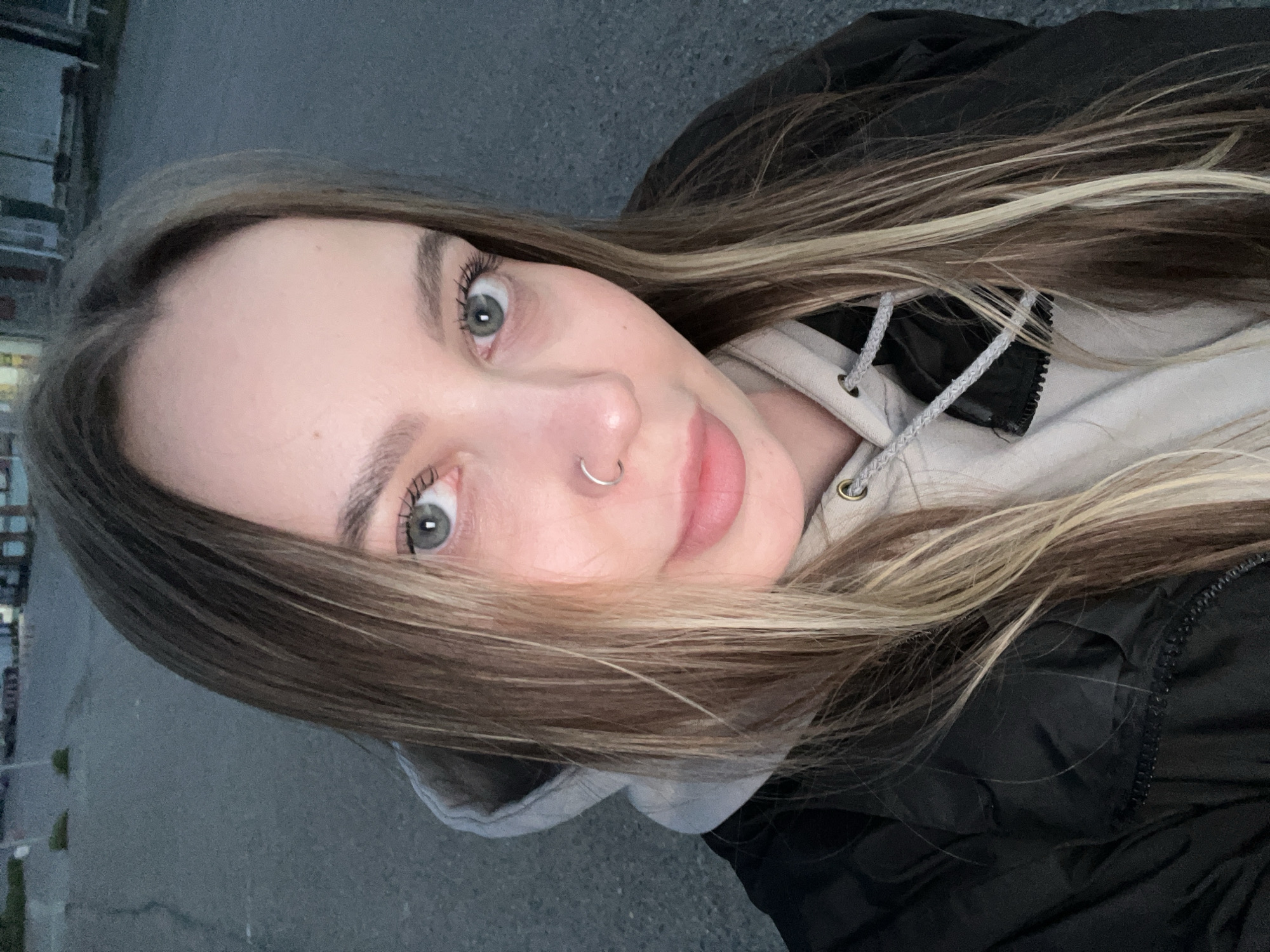 A young woman is featured in a close-up, upward-angled selfie, looking directly into the camera with wide, light green eyes and a subtle, closed-mouth smile. Her long, straight hair, which is a blend of brown and blonde shades, falls around her face and shoulders. A silver hoop nose ring adorns her left nostril. She is wearing a light-colored (possibly beige or light grey) hooded sweatshirt under a dark, likely black, zippered jacket, with the hood drawstrings visible. The background shows an outdoor setting with a dark, paved surface, resembling asphalt or concrete. Blurred structural elements are faintly visible in the distance, indicating an urban or suburban environment. The lighting is soft and diffused, suggesting either an overcast day or twilight. This moment was captured in Novopokrovskaya, Russia. No visible text is present in the image.