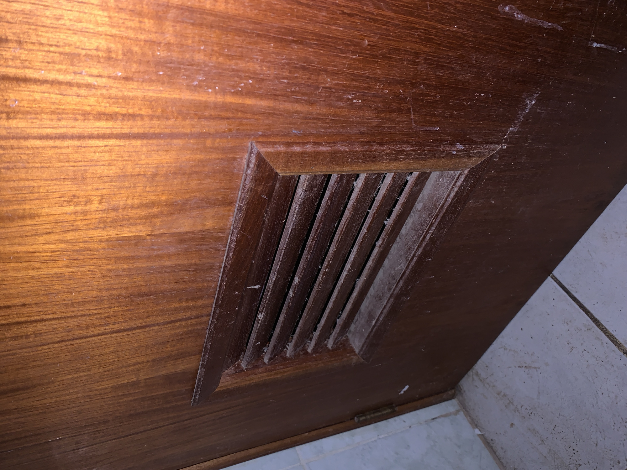 The image shows a brown wooden door with a built-in ventilation grille. The grille is rectangular and has horizontal slats. Both the door and the grille appear dusty and have some stains. The lower right corner of the image shows a glimpse of white tiles, likely part of a floor or wall. There is no information in the image to suggest this is in Santa Ana, Venezuela.